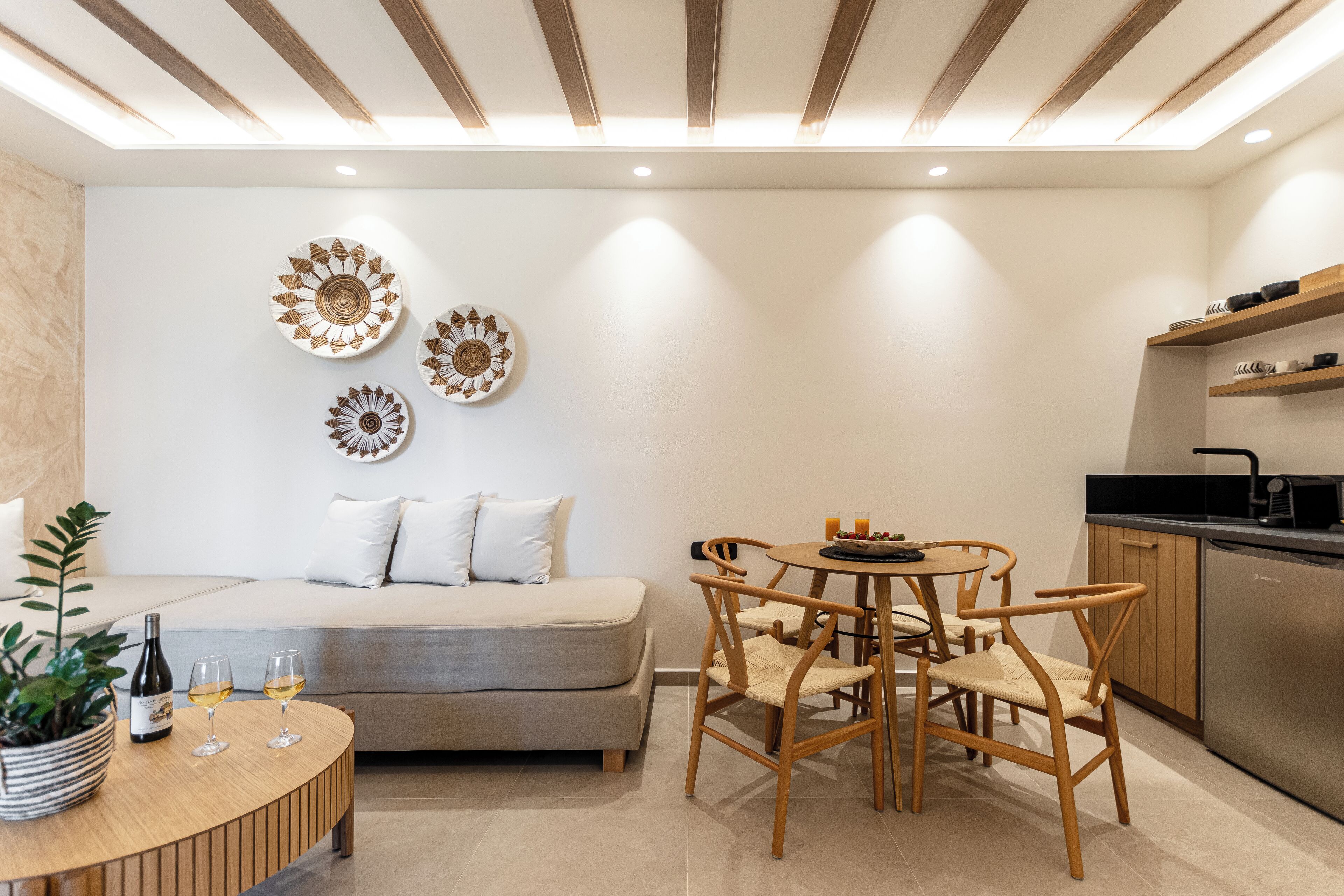 Uma sala de estar moderna com um sofá, uma mesa redonda de madeira e uma área de cozinha com máquina de lavar louça e prateleiras.