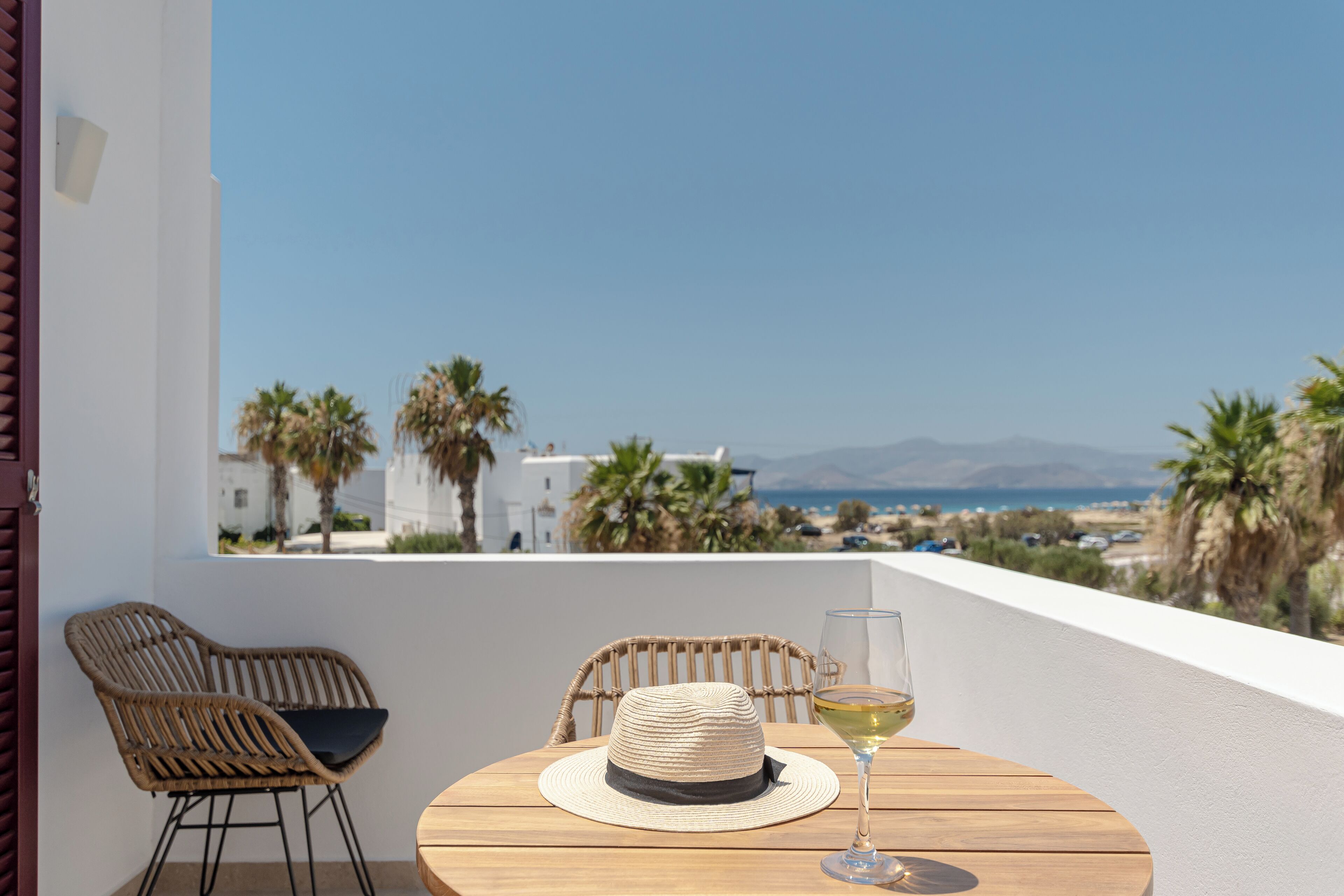 A balcony with a table, two wicker chairs, a straw hat, and a glass of white wine.