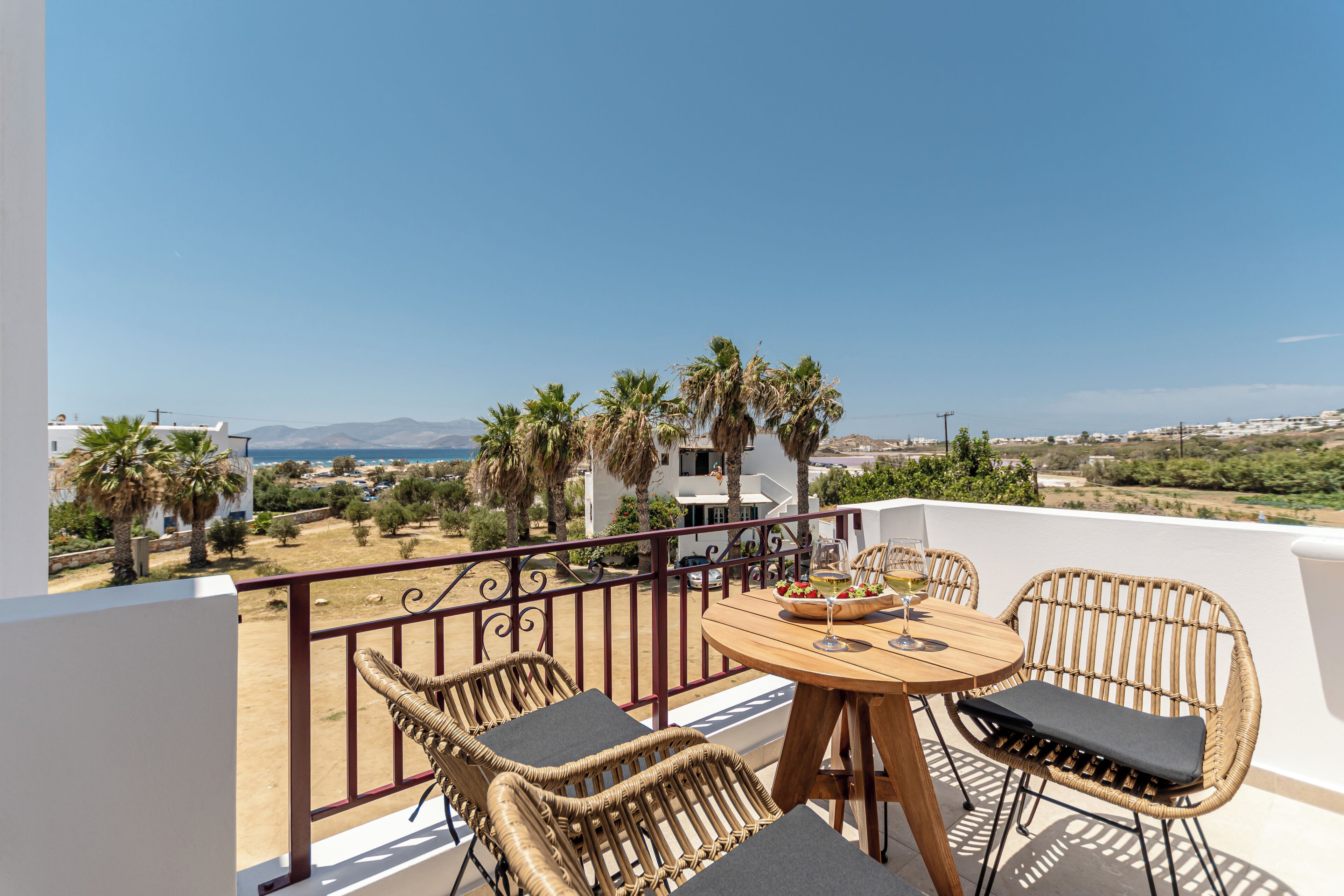 A balcony with wicker chairs and a table, overlooking a coastal landscape with palm trees and buildings.