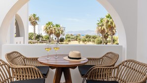 Mesa redonda con sillas de mimbre y dos copas de vino blanco, frente a una pared blanca con vistas a palmeras y montañas.