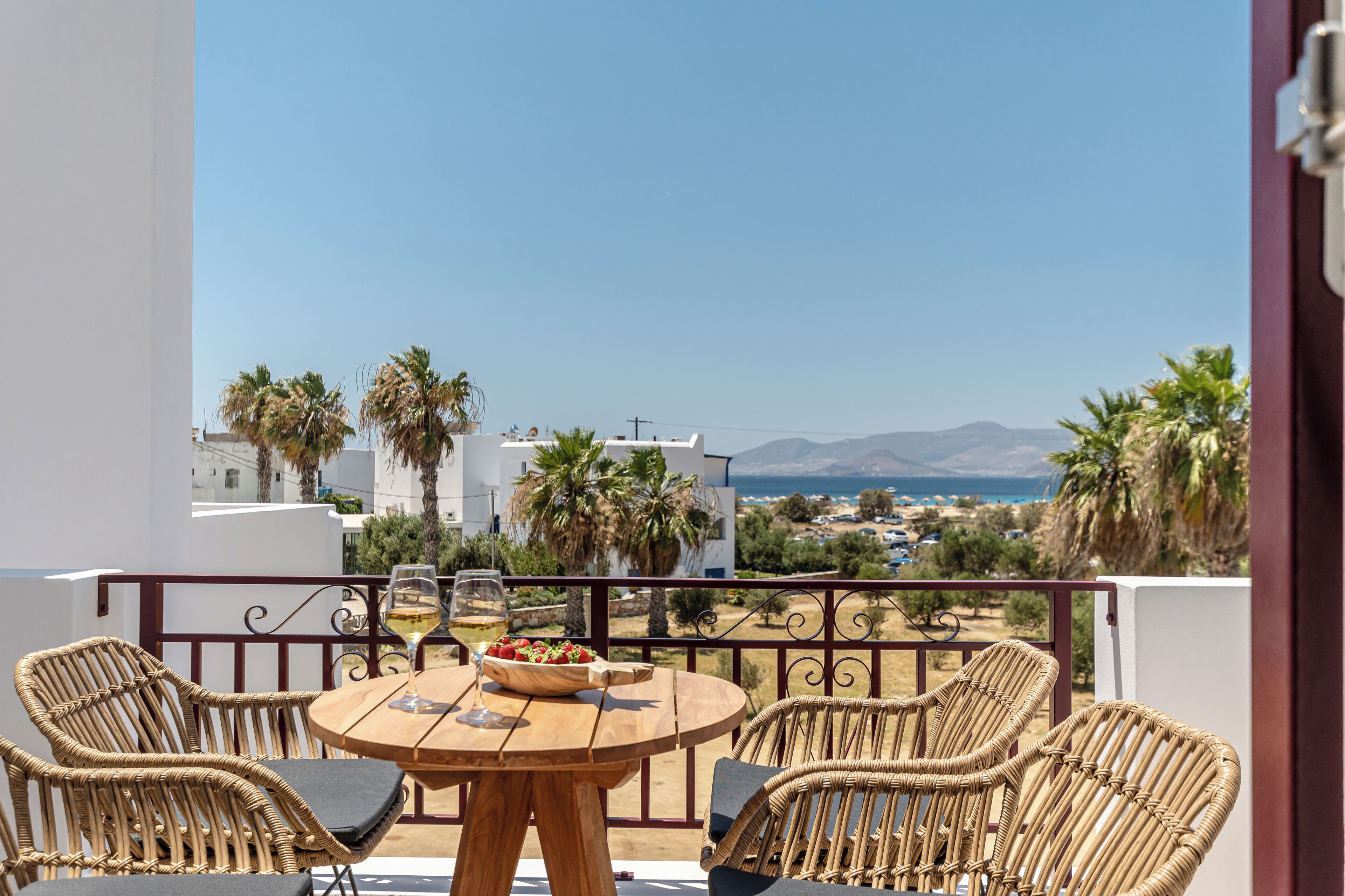 Un balcón con mobiliario de mimbre, una mesa con un cuenco de fruta y copas de vino, y vistas al mar y edificios.