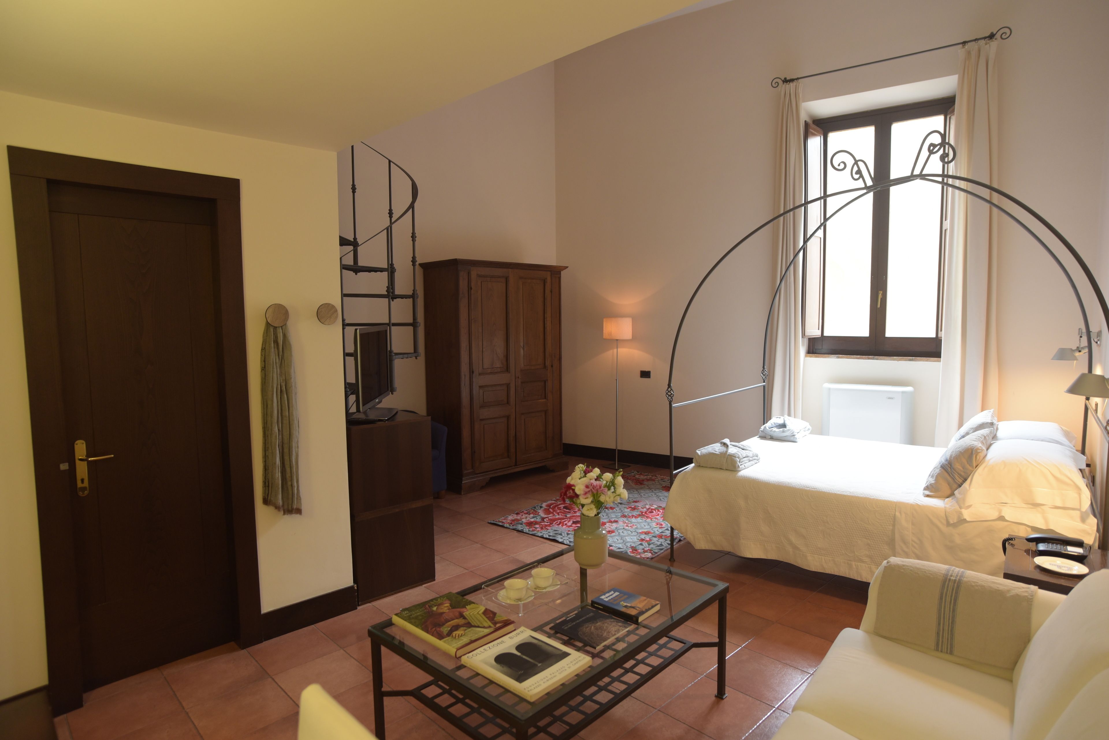 A cozy room with a bed, a small table with books and a vase, a wooden cabinet, and a window with curtains.