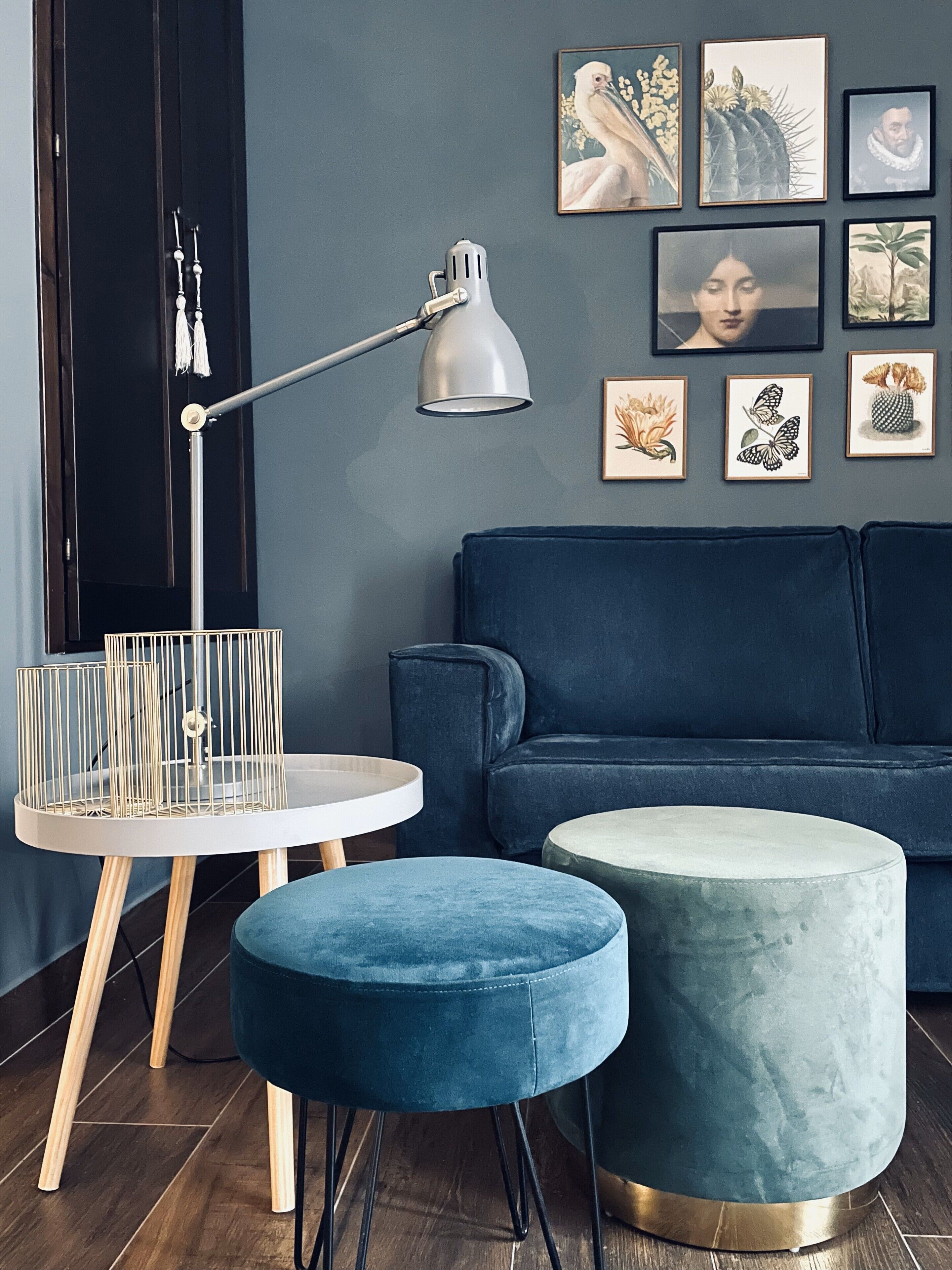 Un salón moderno con un sofá azul oscuro, una mesa redonda con tapa blanca, un otoman azul y una lámpara de pie. En la pared hay cuadros enmarcados.