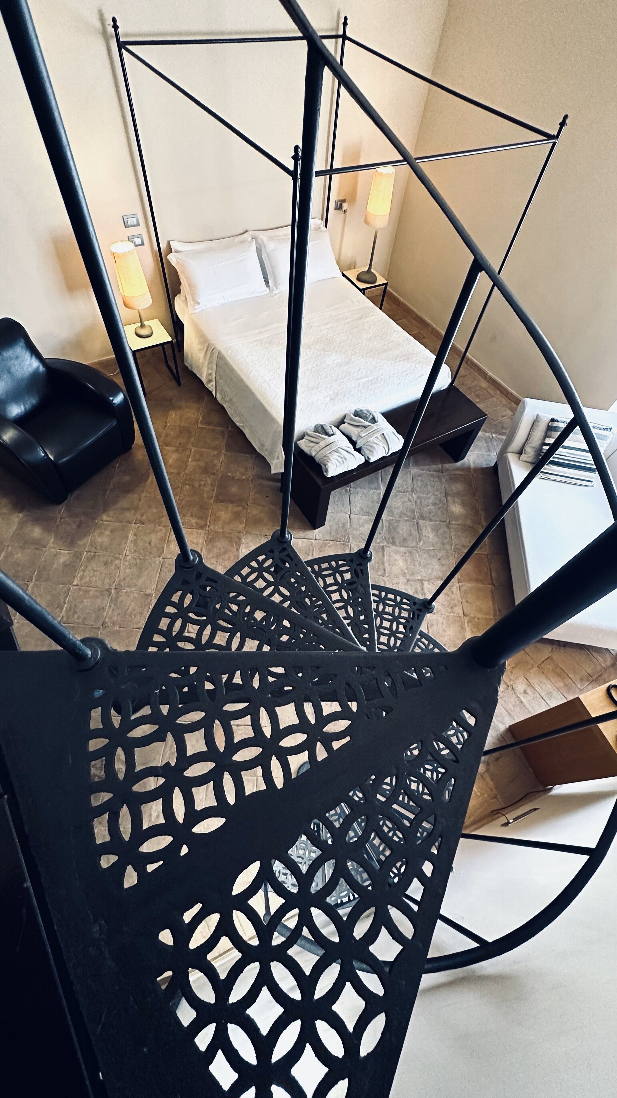 Una escalera de caracol con un diseño metálico geométrico, que conduce a un dormitorio con cama, mesitas de noche y una lámpara.