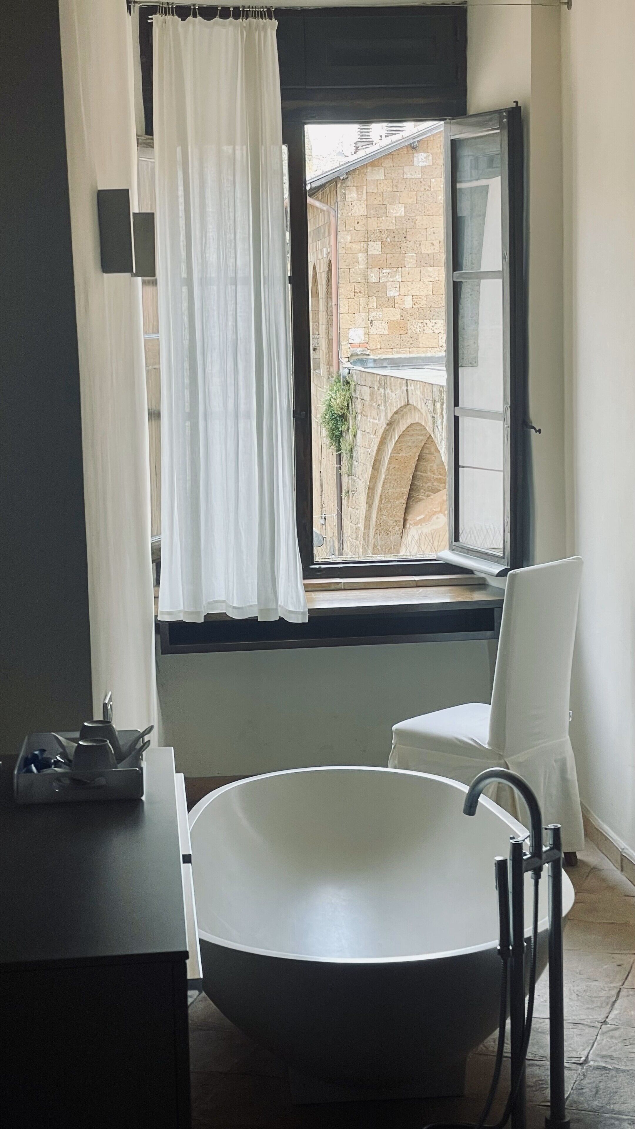 A bathroom with a freestanding bathtub, a window with a view of a brick building, and a chair.