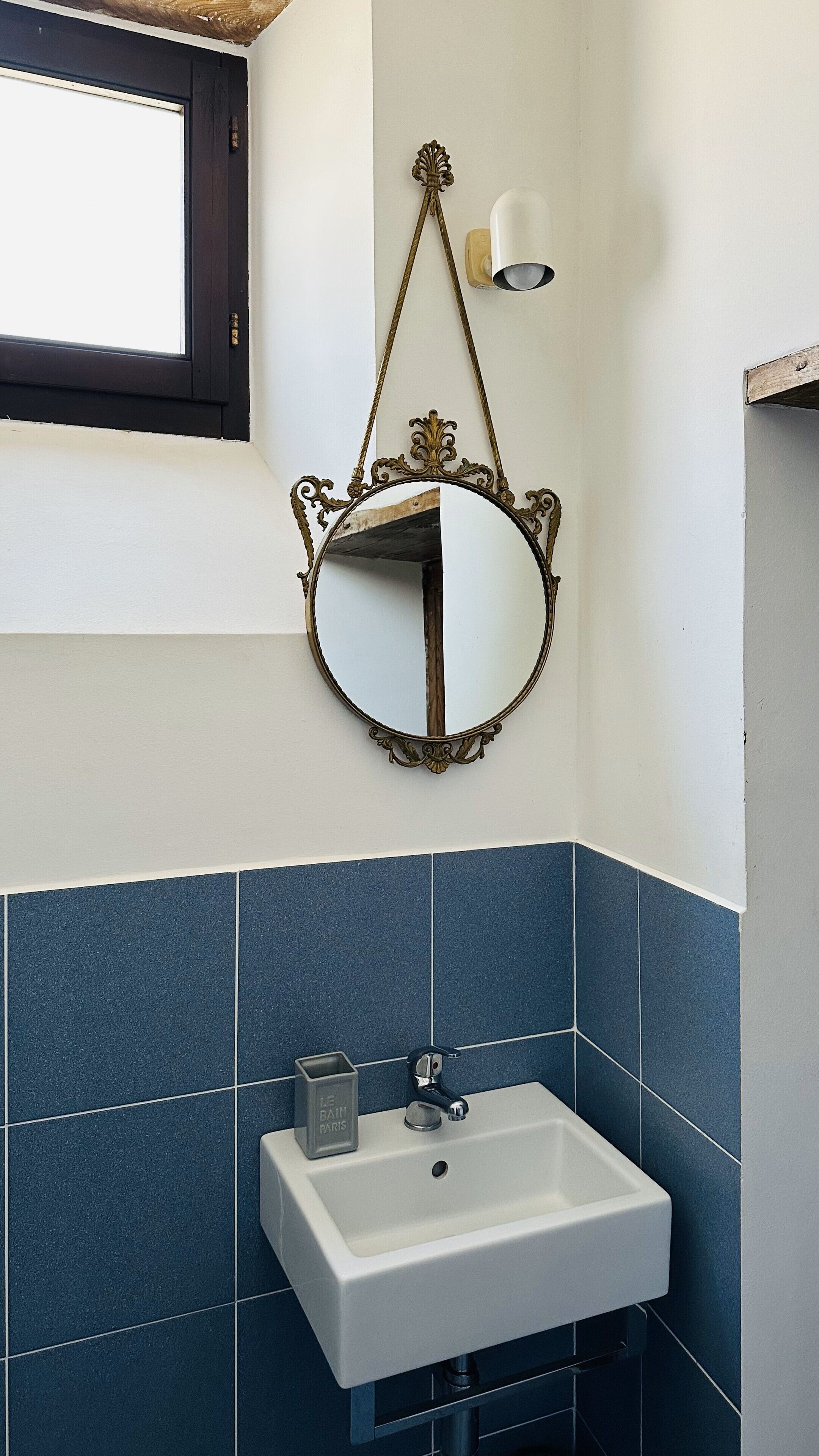 A bathroom with a white sink, blue tiles, a decorative mirror with an ornate frame, and a wall-mounted light fixture.