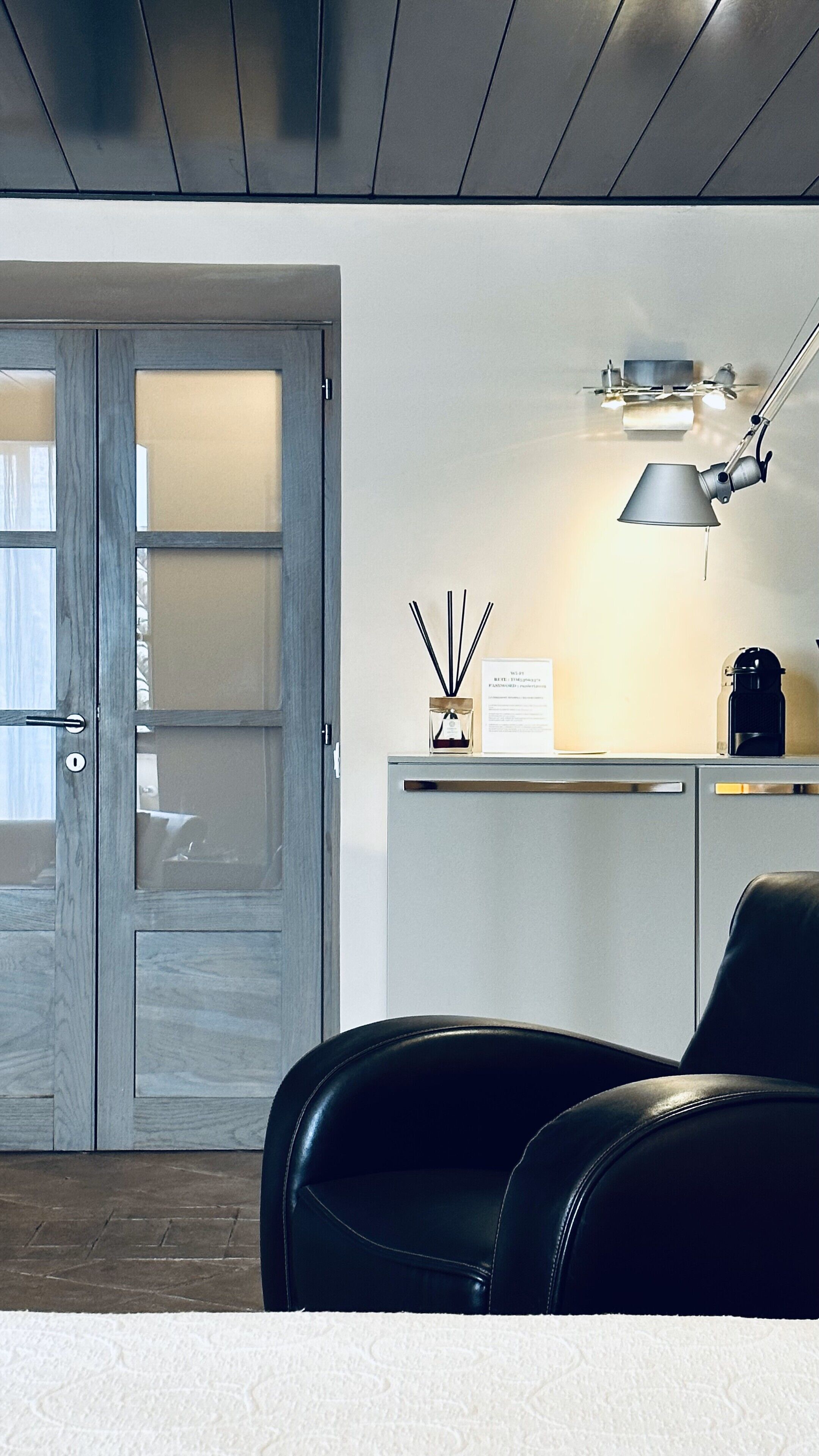 Ein modernes Zimmer mit einer Holztür, einem Schreibtisch mit Lampe, einem Stuhl und einem kleinen Tisch mit Kerze und Schild.