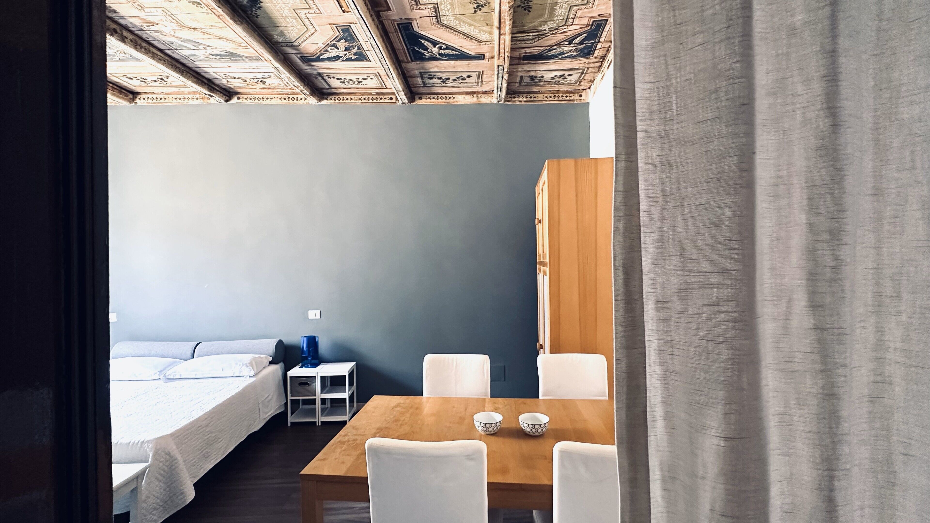 A room with a bed, a table with two chairs, a small white cabinet, and a wooden wardrobe.