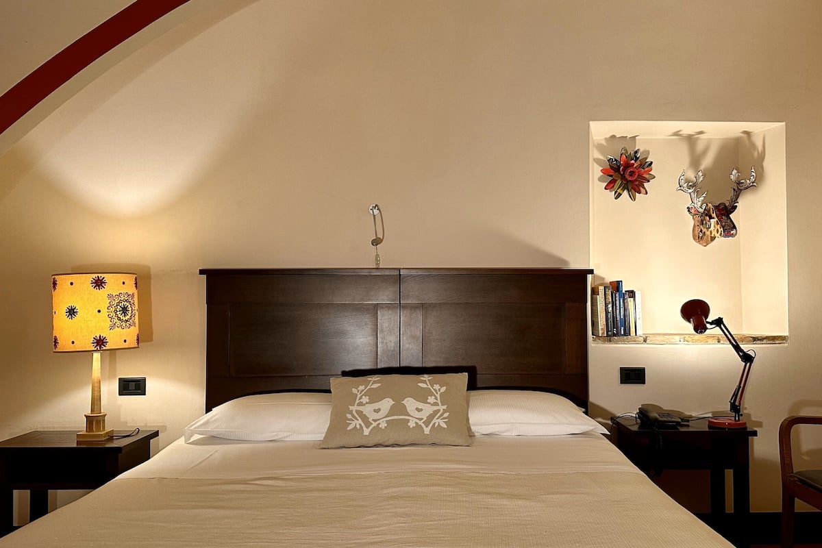 Ein Schlafzimmer mit einem großen Bett, einem Nachttisch mit Lampe, einem Sessel, einem Schreibtisch mit Telefon und einem Fenster mit Dekoration.