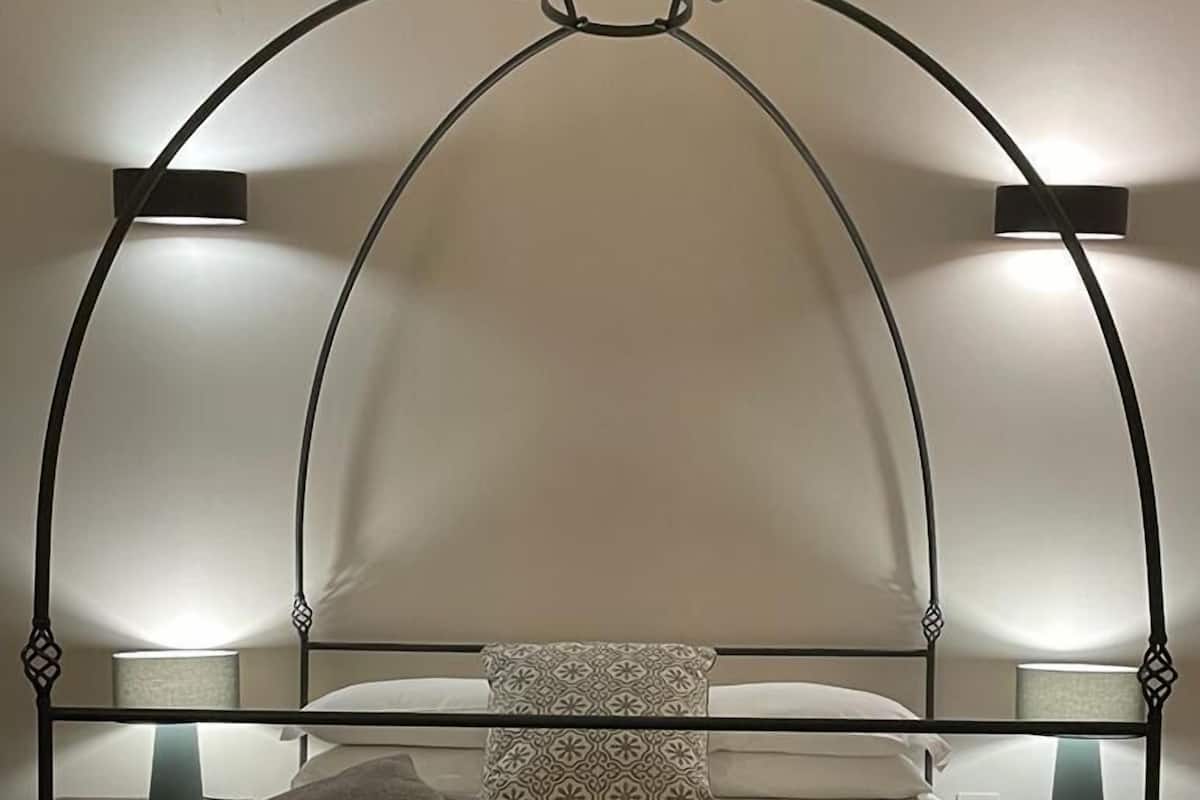Ein Bett mit Metallgestell, zwei Nachttischlampen, ein gemustertes Kopfkissen und gefaltete Handtücher auf dem Nachttisch.