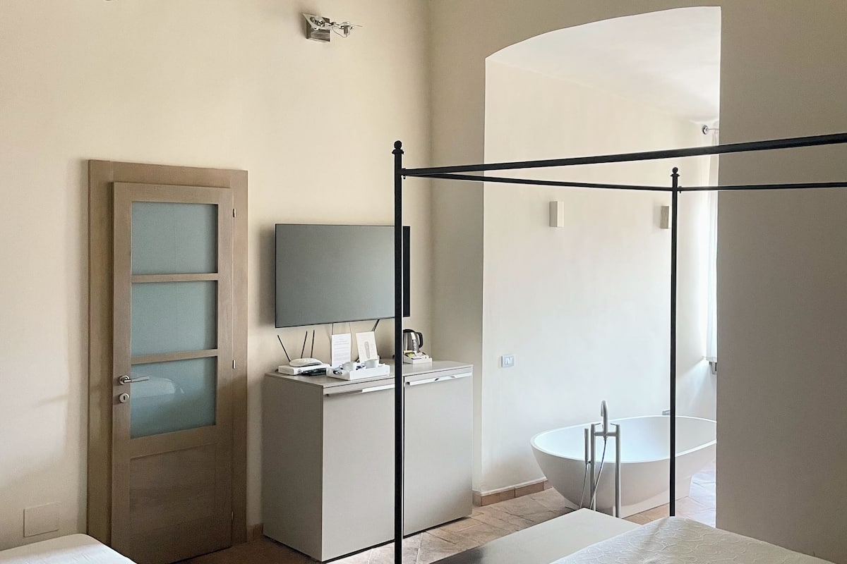 Ein Schlafzimmer mit Himmelbett, einer Holztür mit Sandstrahlglaseinsätzen, einem Nachttisch mit Lampe und Blick ins Badezimmer.