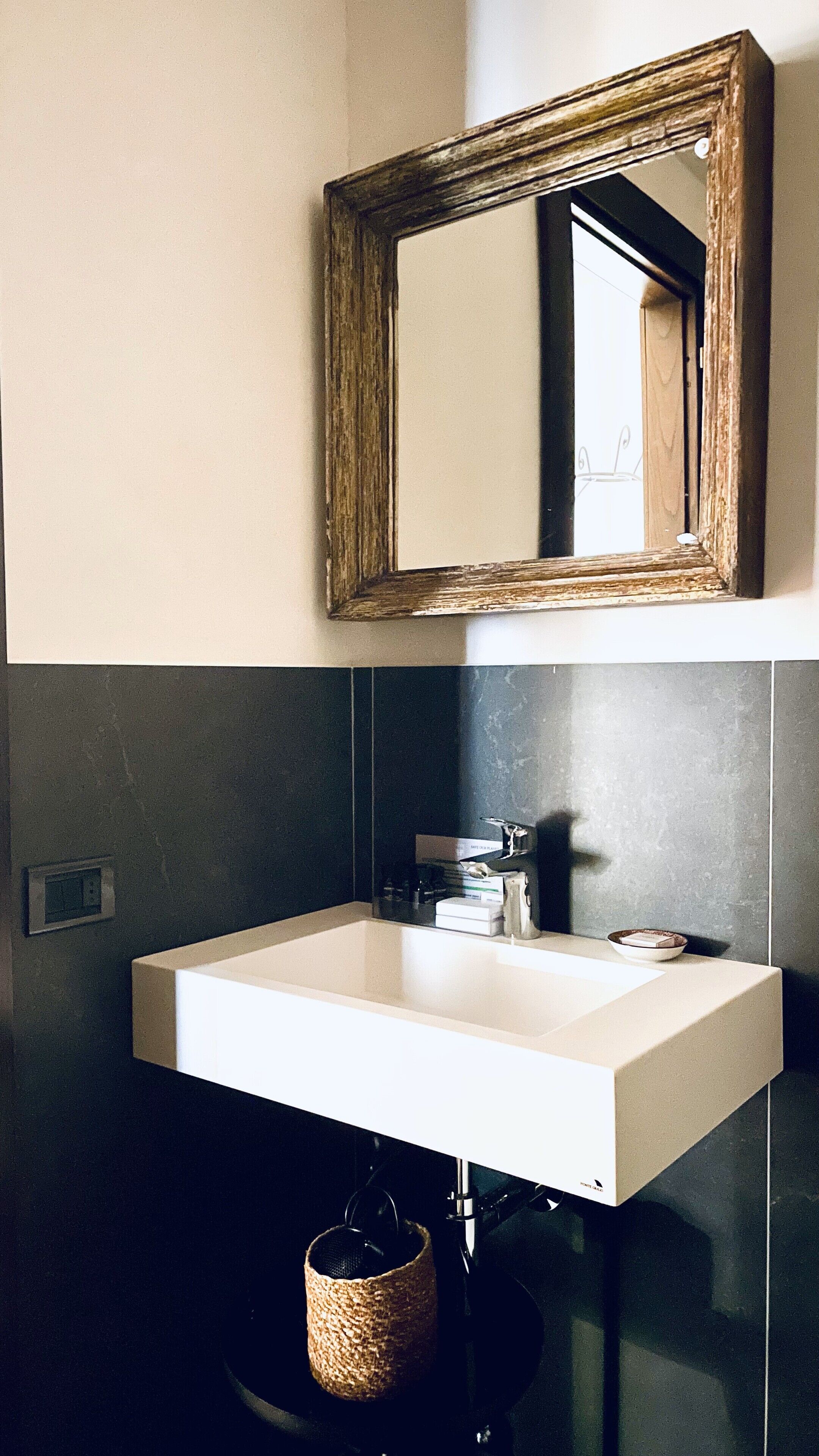 A bathroom with a white sink, a wooden-framed mirror, and dark tiled walls.
