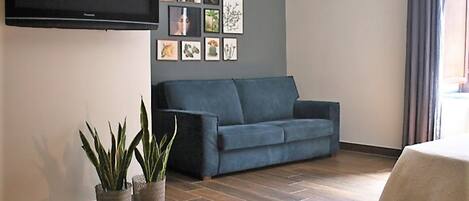 A living room with a dark blue sofa, a flat-screen TV mounted on the wall, and several framed botanical prints. There are two potted plants on the floor.