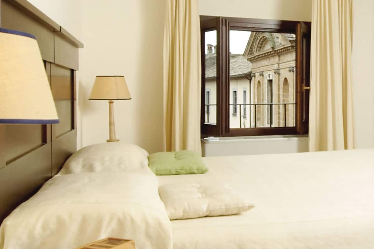 Ein Hotelzimmer mit Bett, Nachttisch mit Lampe und Fenster mit Blick auf ein Gebäude.