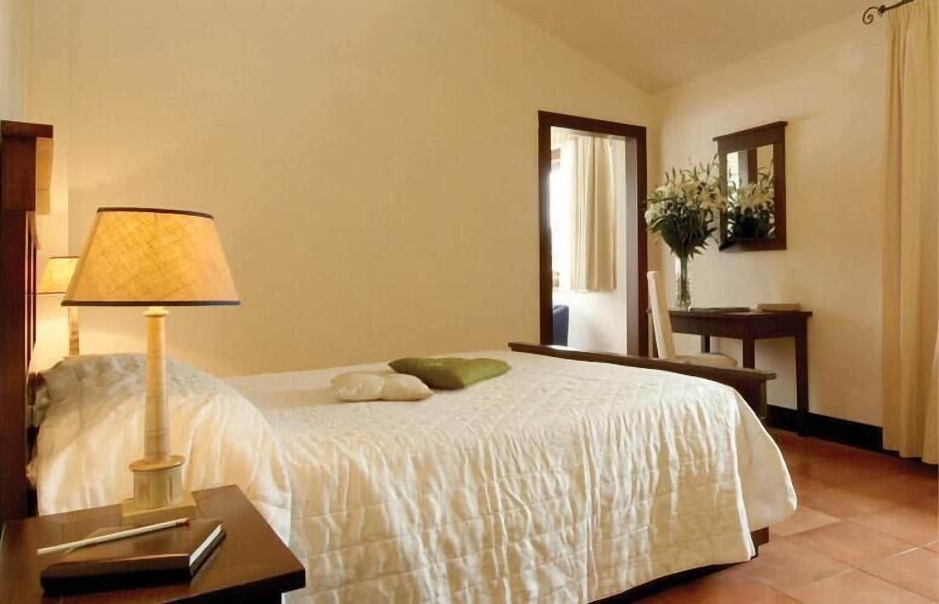A hotel room with a bed, a nightstand with a lamp, a desk with a mirror, and a vase with flowers.