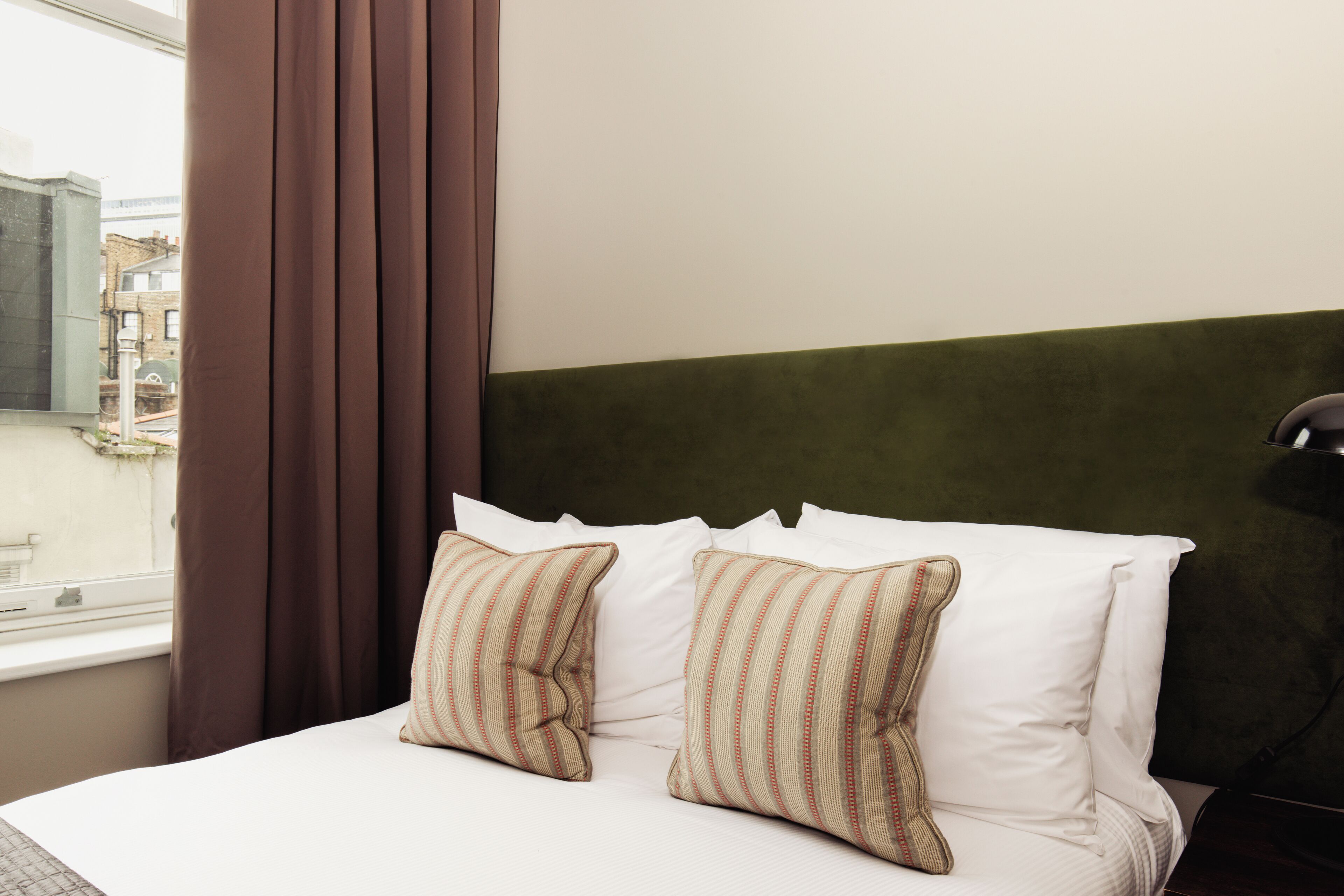 A hotel room with a bed, two pillows, a window with curtains, and a view of buildings outside.