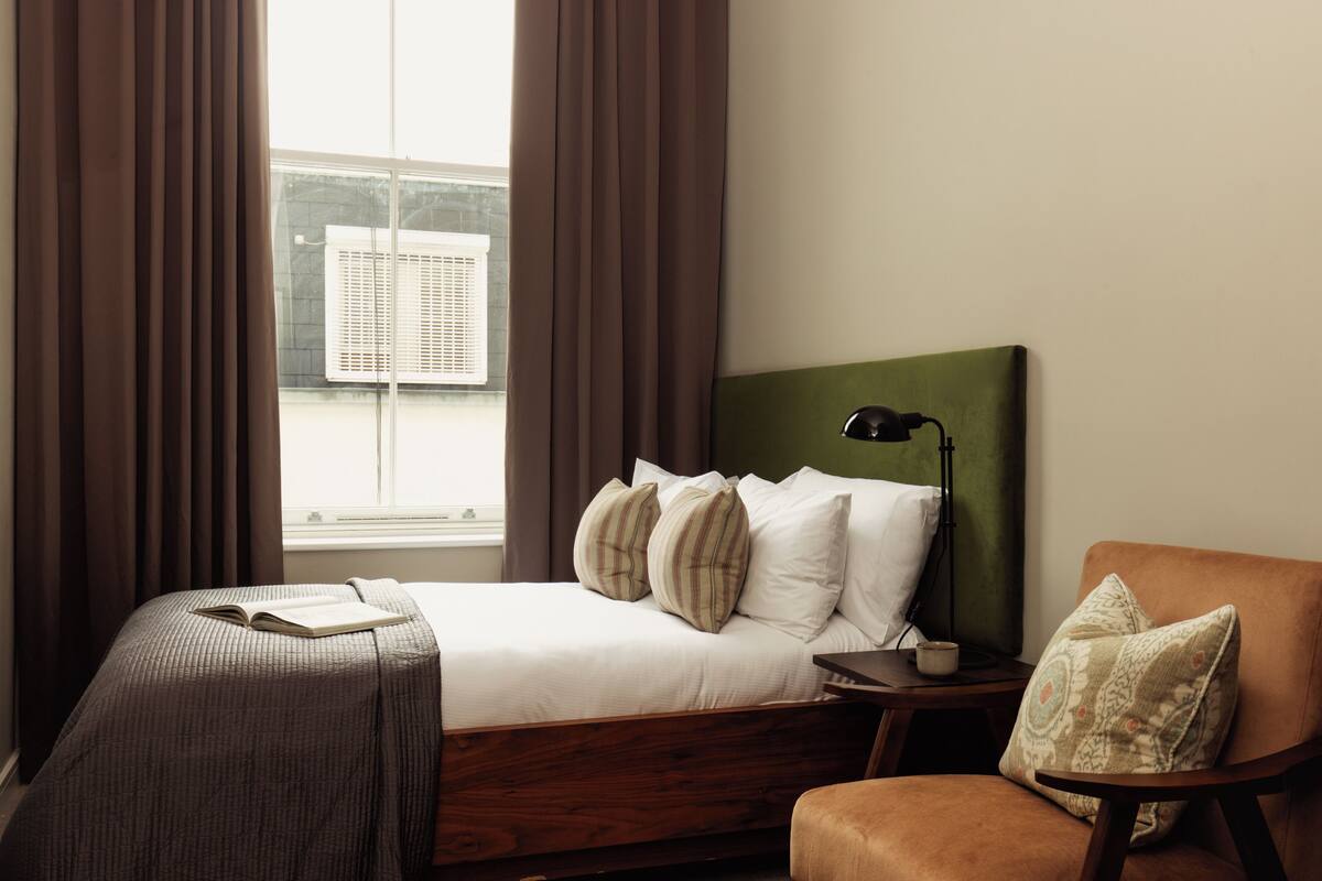 Una cama bien hecha con almohadas, una mesita de noche con una lámpara, una silla y una ventana con cortinas.