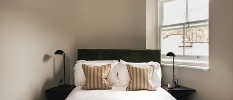 A neatly made bed with striped pillows, a bedside table with a lamp, and a window with a view of buildings.