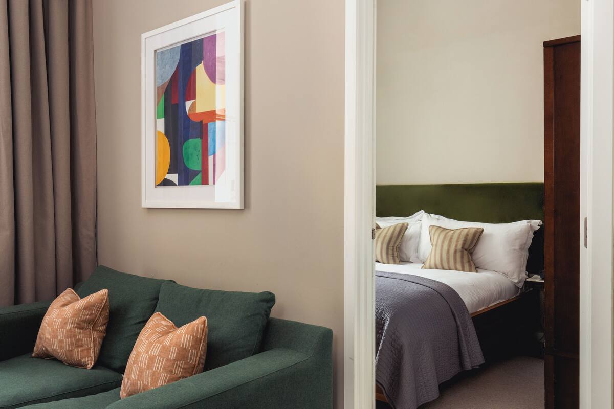 Una habitación con un sofá verde, un cuadro abstracto enmarco y una cama con cabecera verde.