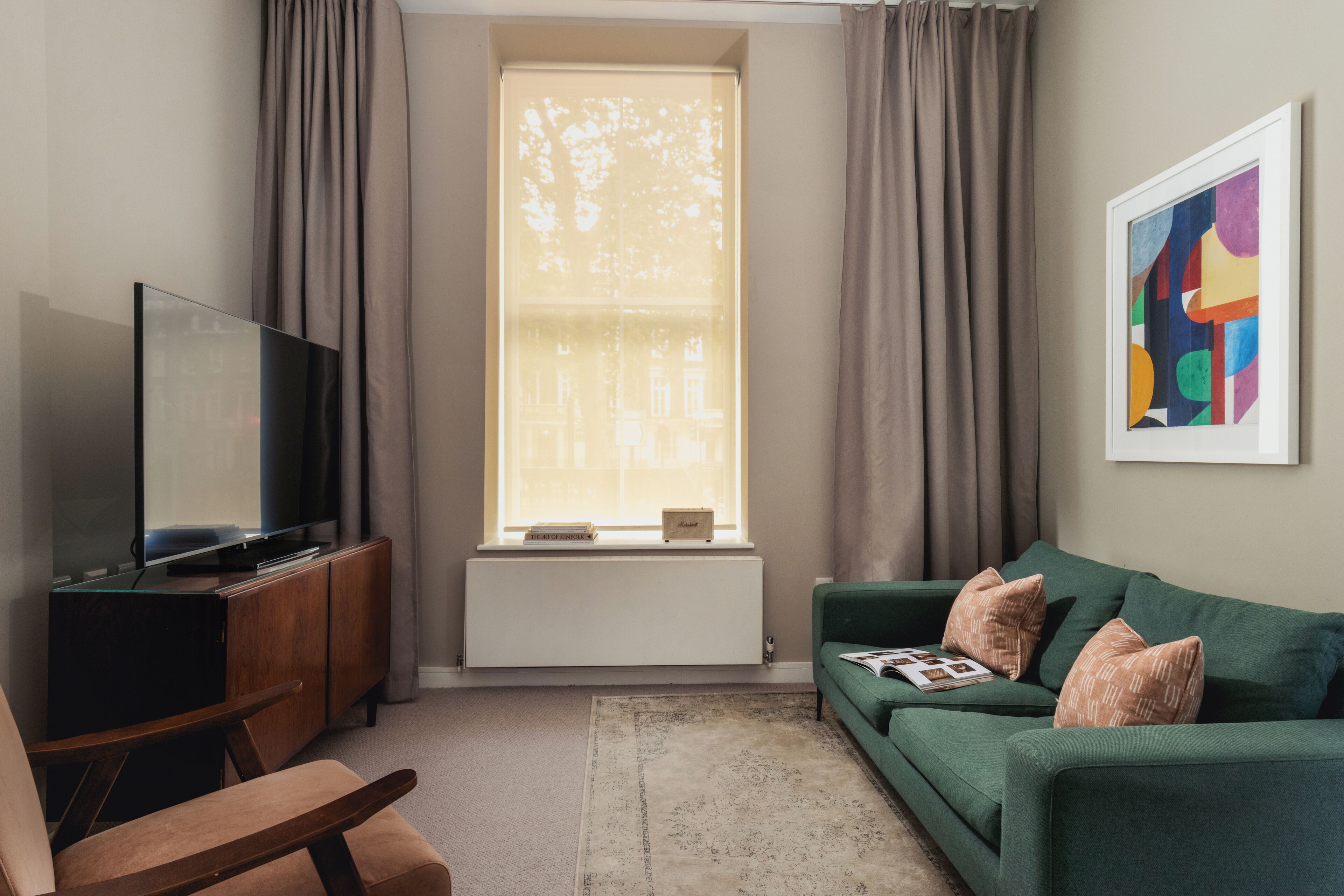 Ein Wohnzimmer mit Fernseher, einer Couch, einem Sessel, einer Heizung und einem fenster mit Vorhängen.