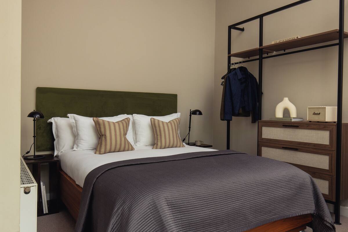 Una cama bien tendida con cabecera verde, una mesita de noche con lámpara, un aparador con espejo y un estante con una chaqueta y un jarrón.