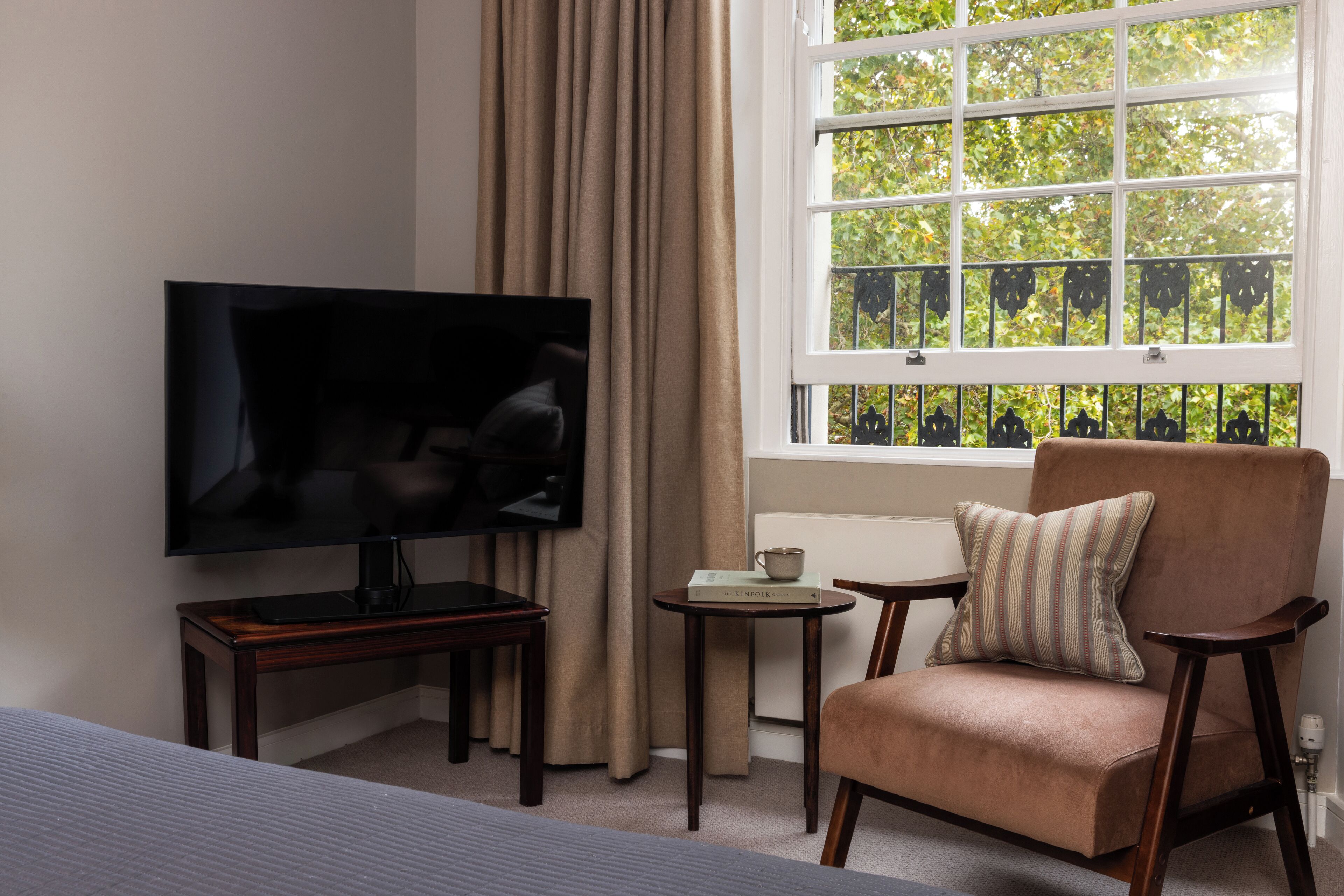 A hotel room with a TV, a chair, a small table, and a window with a view of trees.