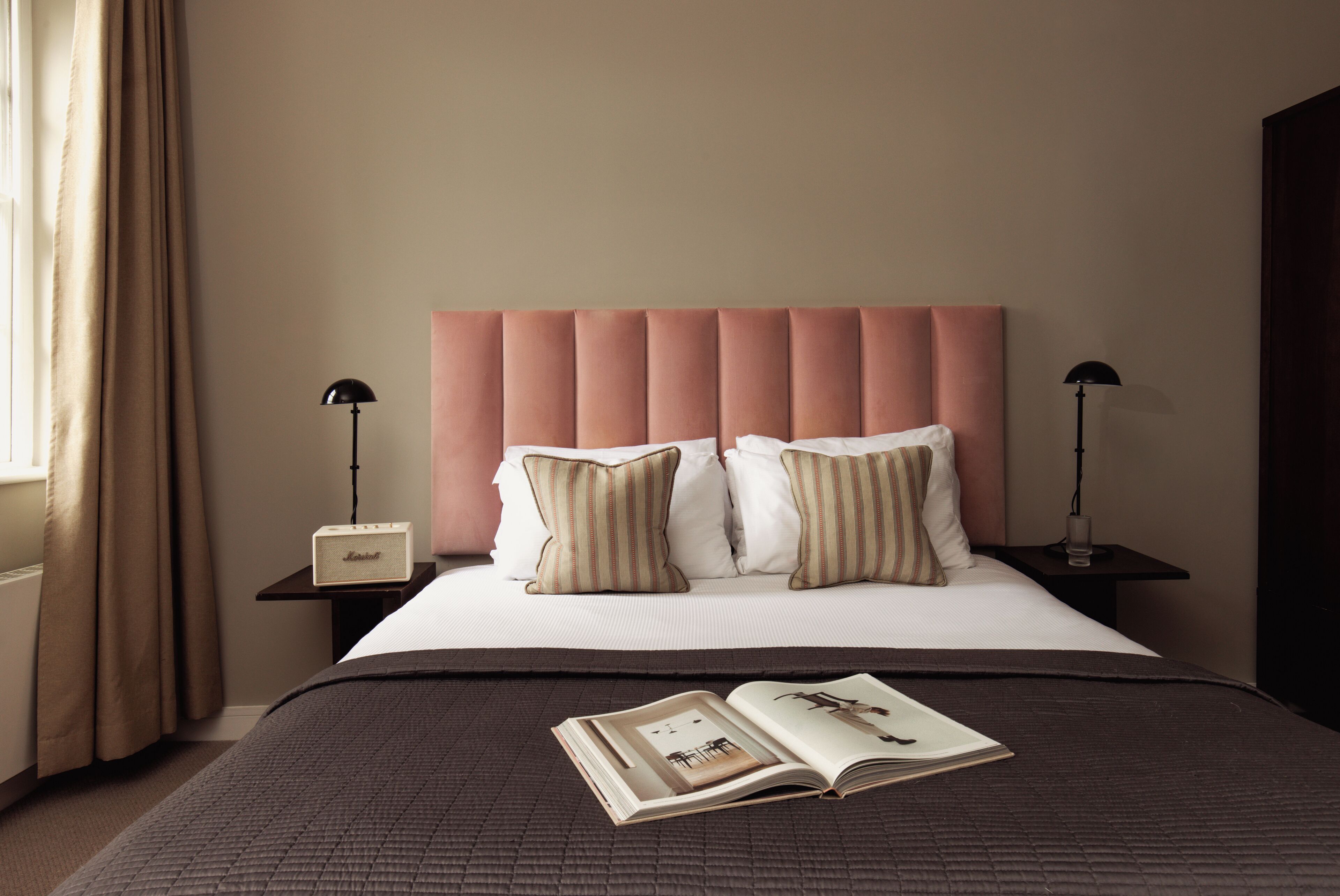 A neatly made bed with a pink headboard, two bedside tables with lamps, and a book on the bed.