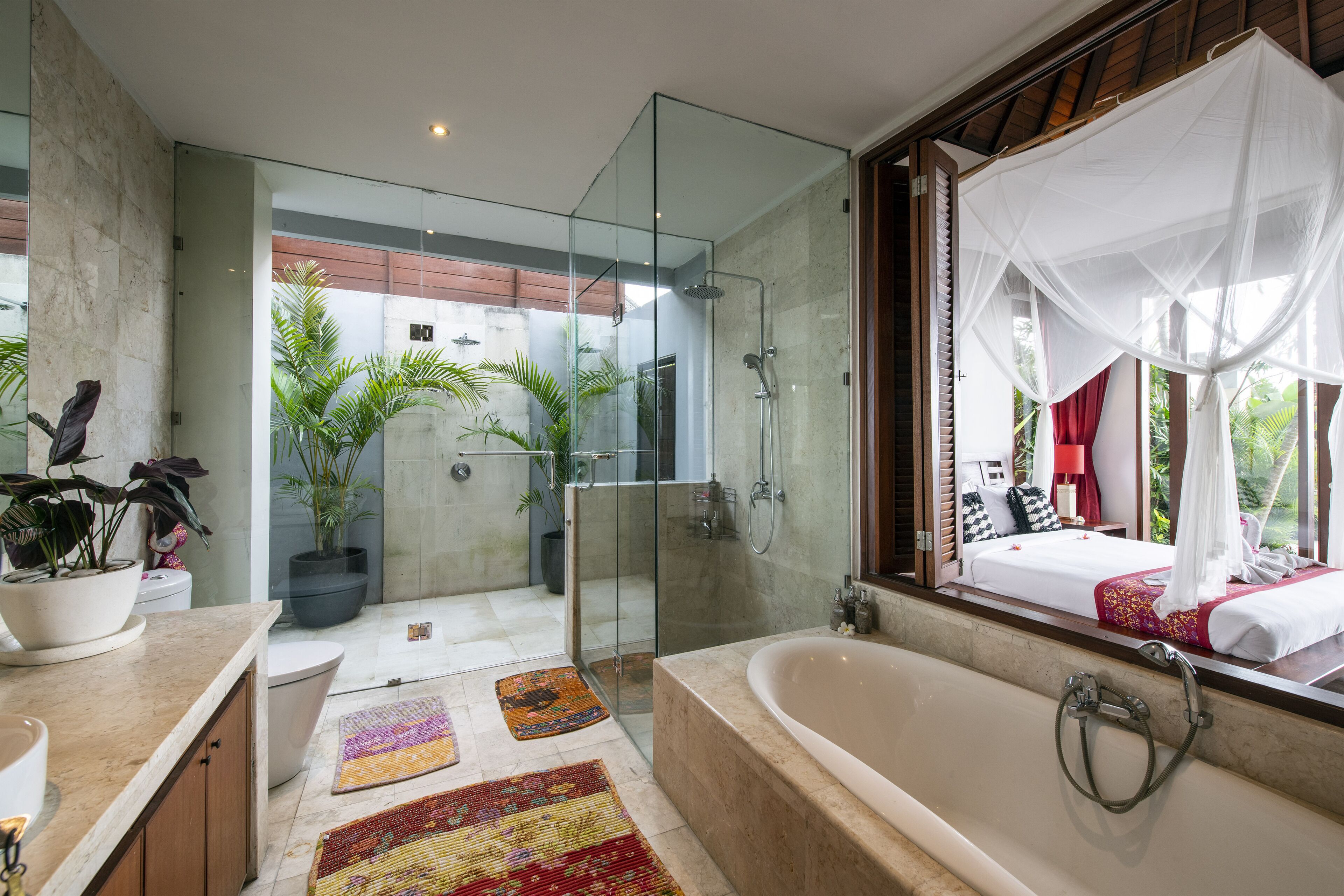 A modern bathroom with a glass-enclosed shower, a large bathtub, and a view of a bedroom with a canopy bed.
