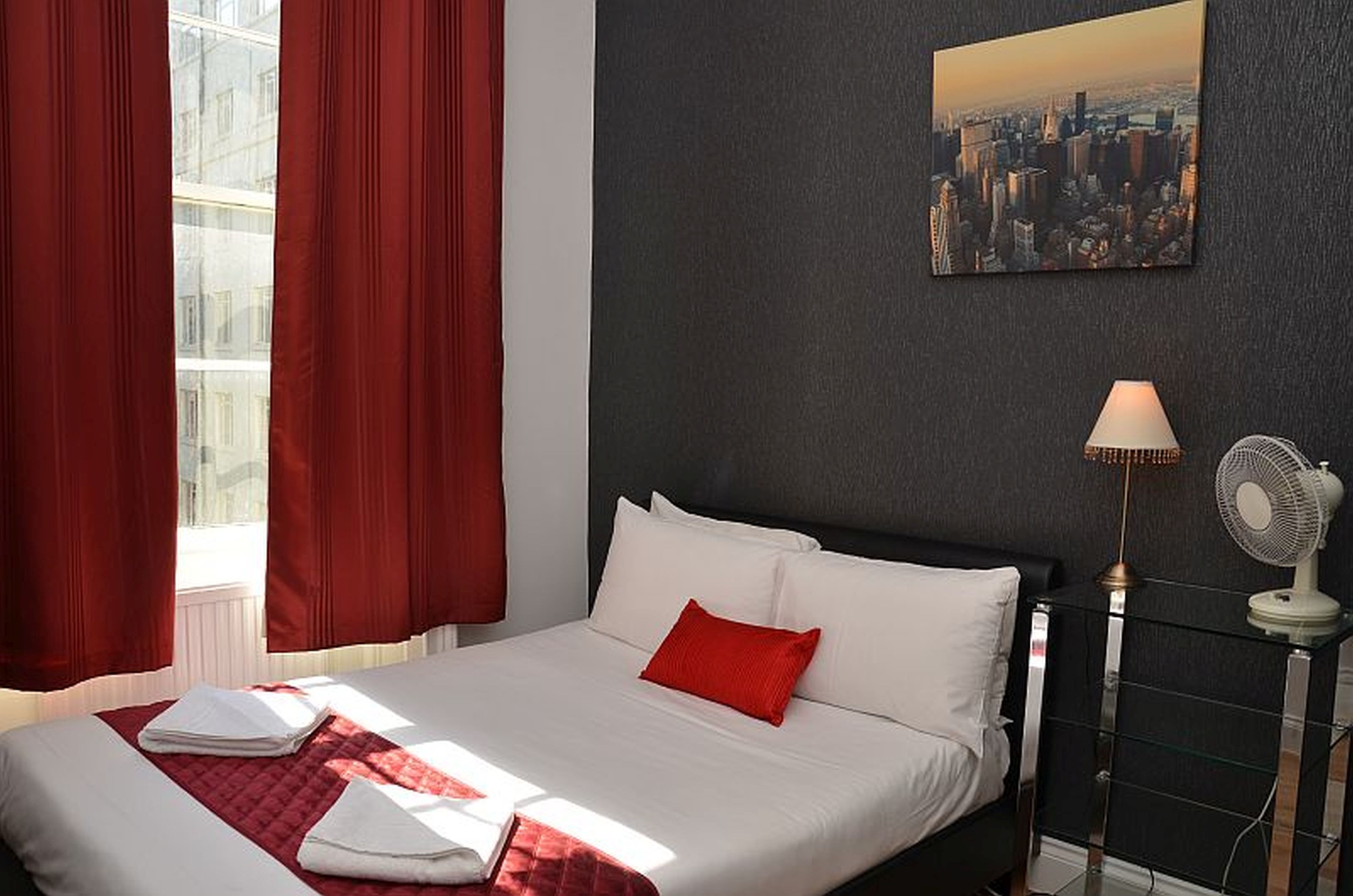 A hotel room with a bed, red curtains, a cityscape painting, a lamp, and a fan.