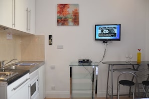 Ett modernt kök med en inbyggd ugn, ett matbord i glas med två stolar och en platt-TV som är monterad på väggen.