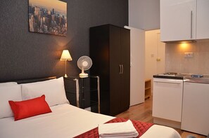 Ett hotellrum med en säng, en röd kudde, en tavla som föreställer en stadsbild, en bordsfläkt, en svart garderob och ett litet kök.