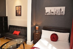 A modern hotel room with a black leather sofa, a glass coffee table, a bed with white and red bedding, a nightstand with a lamp, and a cityscape picture on the wall. - Hyde Park Apartments (London)
