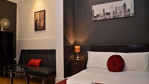 Ein Hotelzimmer mit einem Bett, einem roten Kissen, einem Nachttisch mit Lampe, einer schwarzen Ledersofa und einem kleinen Tisch. An der Wand hängt ein gerahmtes Bild.