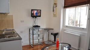 Ein kompakter Wohnbereich mit einer Küche, einer Essecke mit Glastisch, einem an der Wand montierten Fernseher und einem Fenster mit Jalousien.
