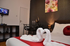 A hotel room with a bed, a TV, a desk with a chair, a small table with two glasses, and a wall painting. - Hyde Park Apartments (London)
