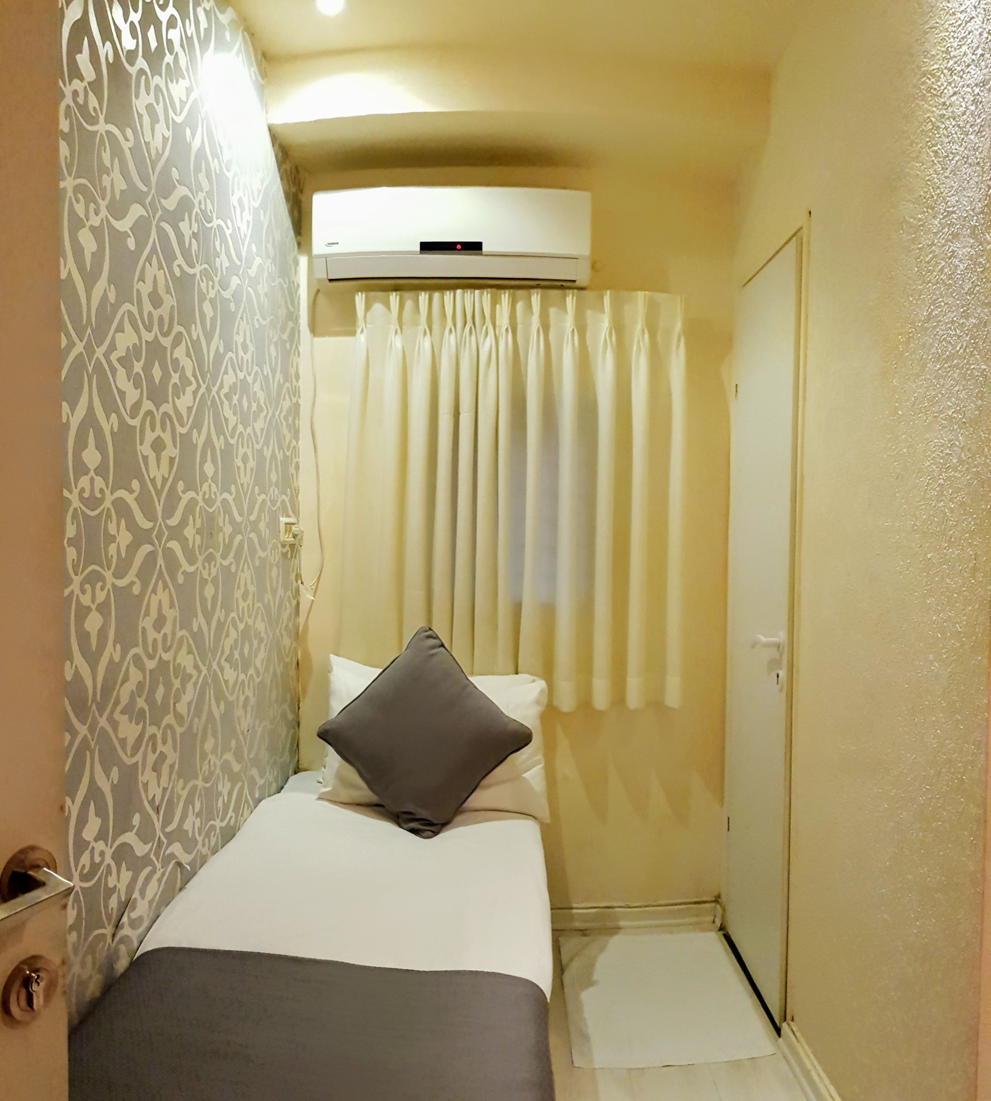 A narrow hotel room with a bed, a pillow, a curtain, and a door.