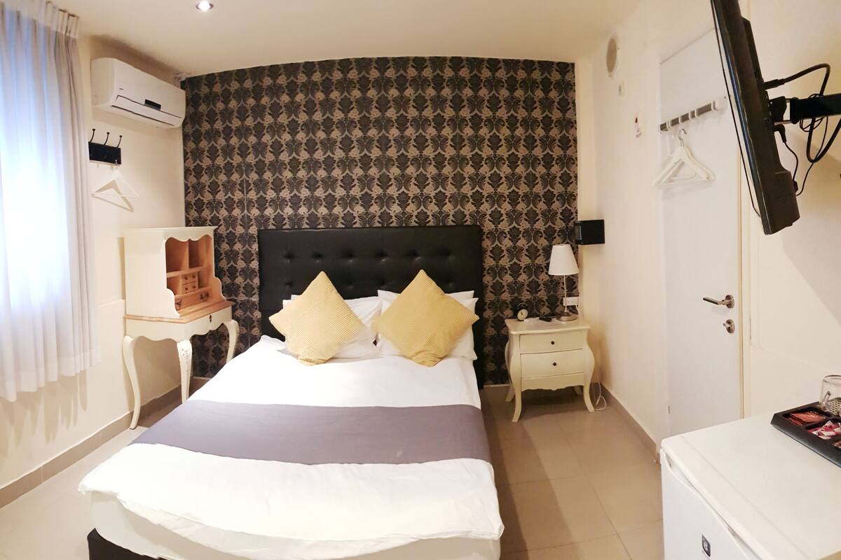 A hotel room with a curved headboard, a bed with white and yellow pillows, a bedside table with a lamp, a wardrobe, and a mirror.
