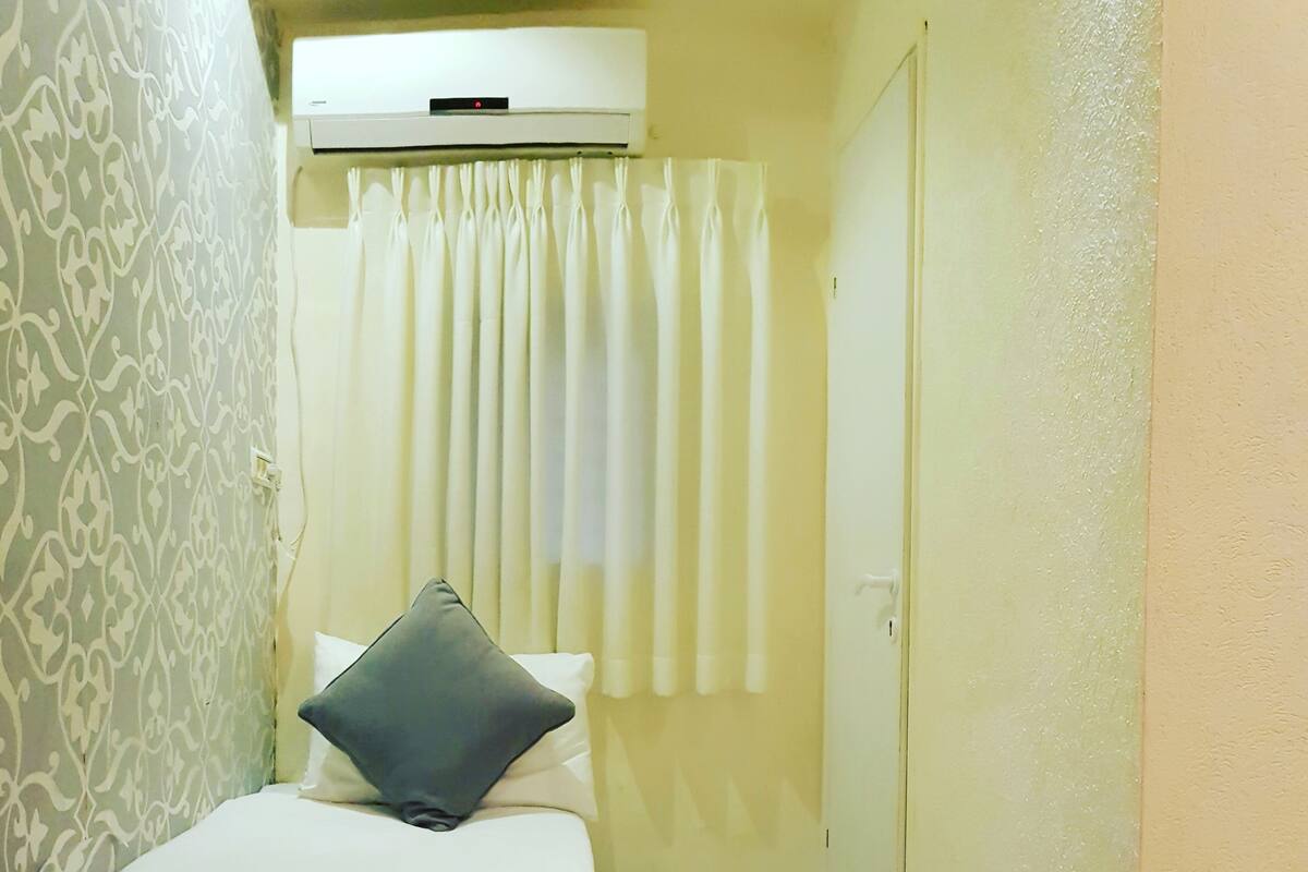 A small, narrow hotel room with a bed, a pillow, a curtain, and an air conditioning unit on the wall.