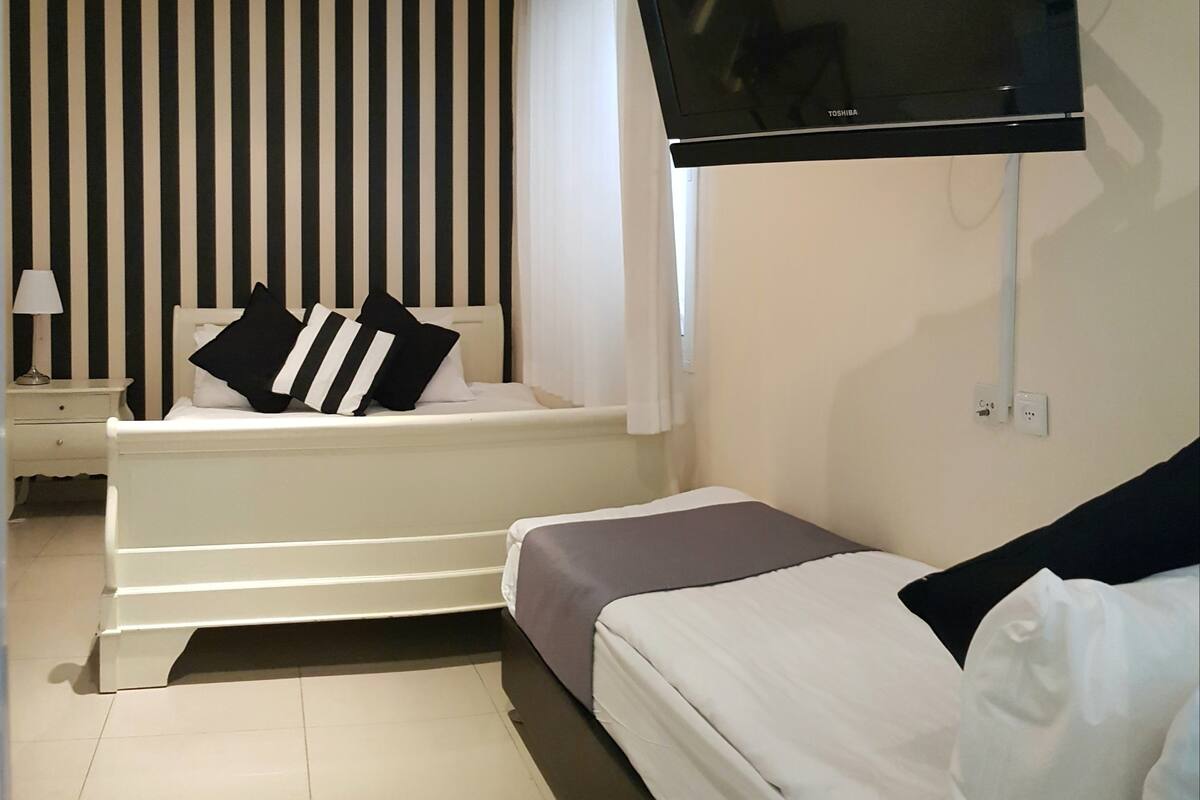 A hotel room with a bed, a sofa, a TV mounted on the wall, a nightstand, and a window with curtains.