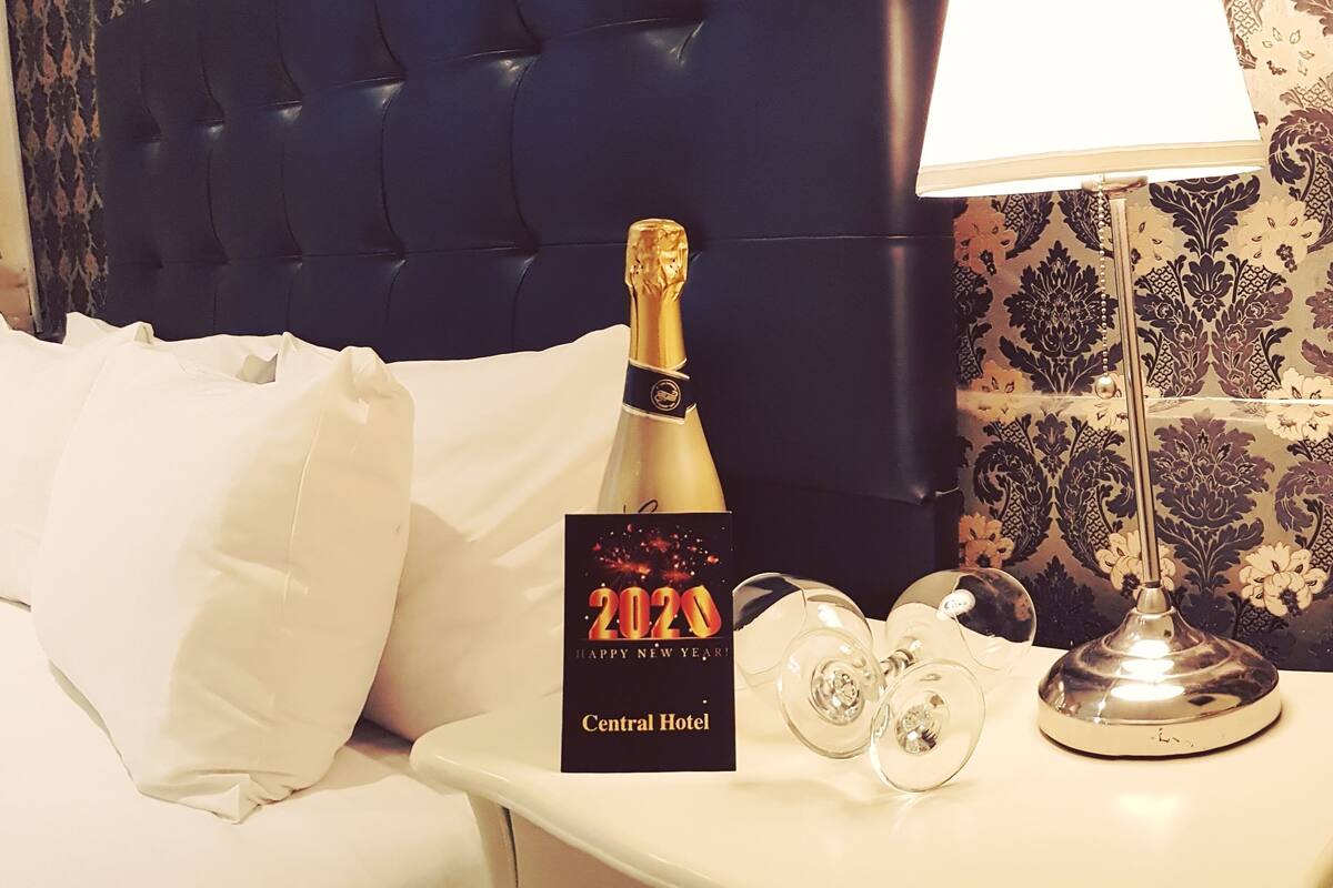 A hotel room with a tufted headboard, a bedside table with a champagne bottle, glasses, and a "2020 Happy New Year" card from Central Hotel.