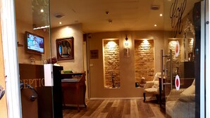 A reception area with a wooden floor, a reception desk, a 'Welcome' mat, and a stone wall with a mirror and a light fixture. - Central Hotel (Tel Aviv)