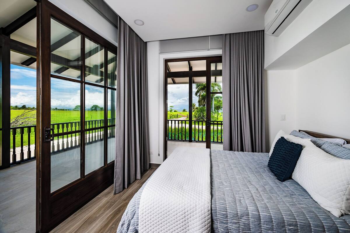 A bedroom with a large bed, a balcony with a view of a green field, and a modern design.