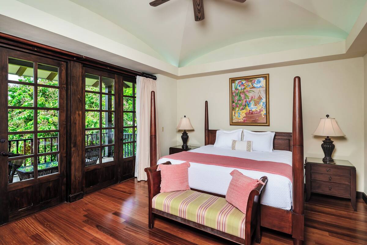A spacious bedroom with a large bed, wooden flooring, a bench, two bedside tables with lamps, and a large window with a view of greenery.