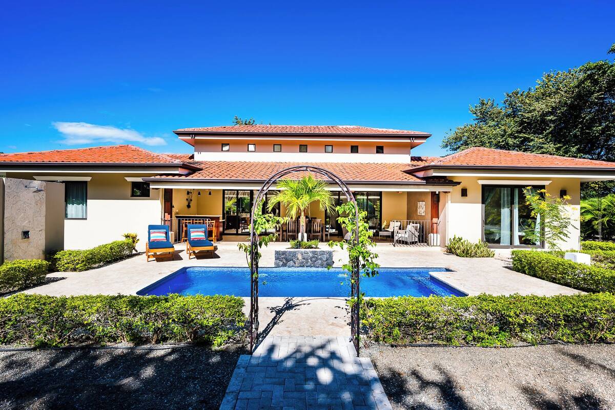 A modern two-story building with a swimming pool in the center, surrounded by well-maintained greenery and a clear blue sky.