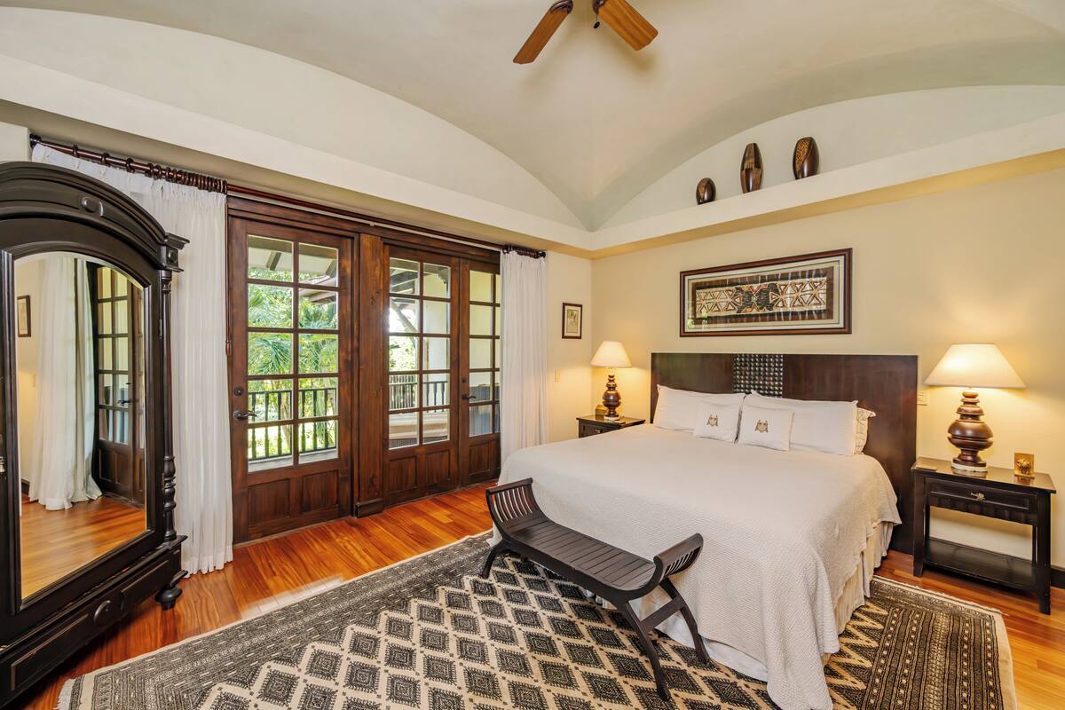 A bedroom with a large bed, two bedside lamps, a wooden bench, a mirror, and a view of greenery through the window.