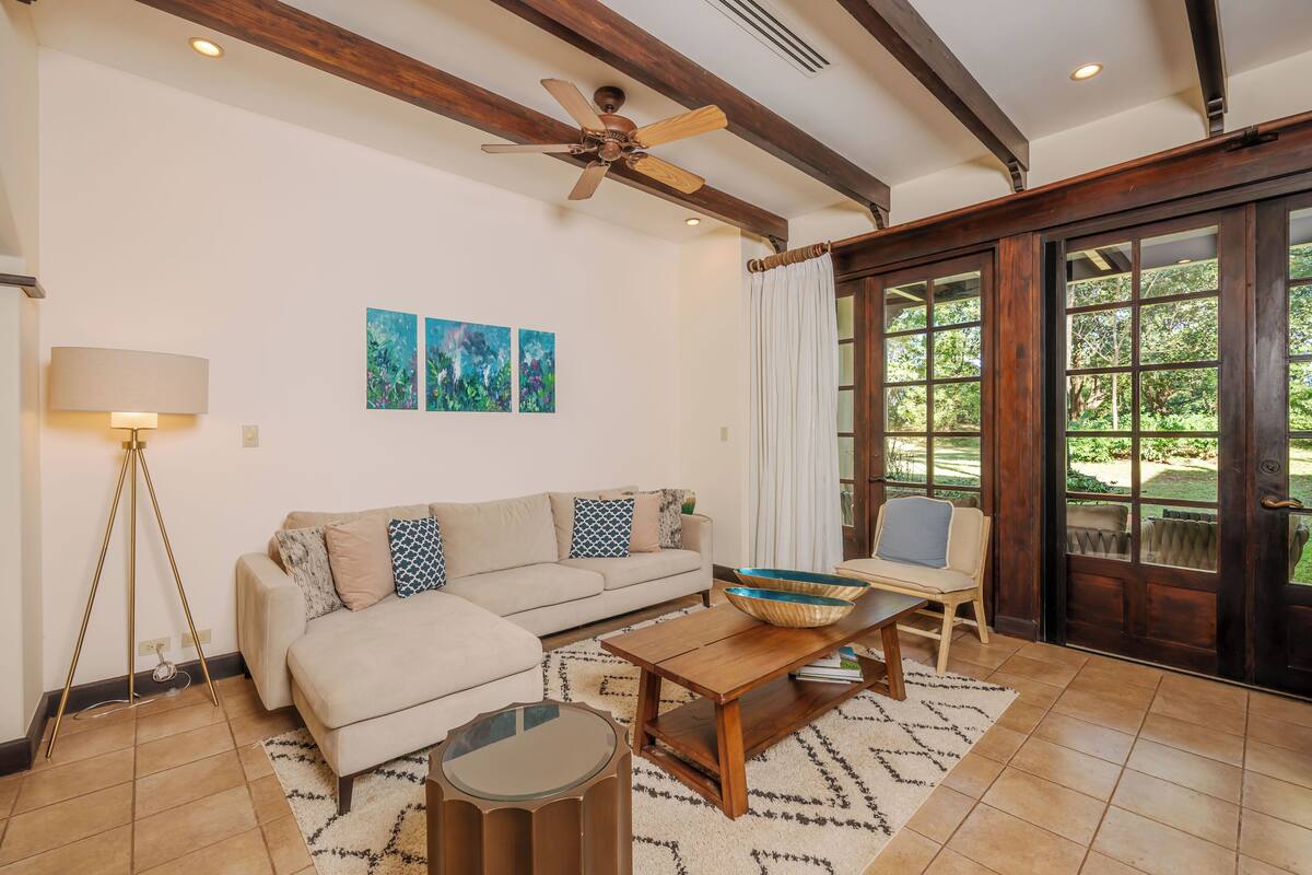 A living room with a sofa, coffee table, floor lamp, and a ceiling fan. There are two paintings on the wall and a sliding glass door leading to an outdoor area.