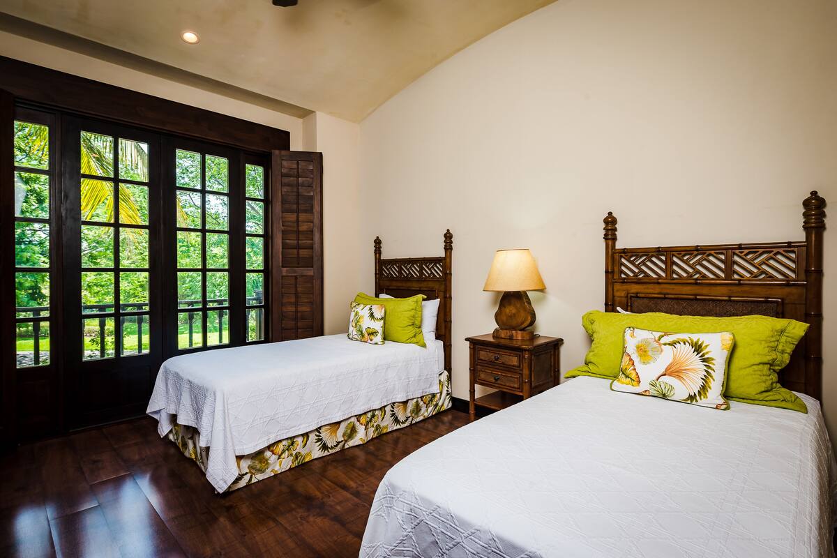 A room with two beds, wooden headboards, and a window with a view of greenery.