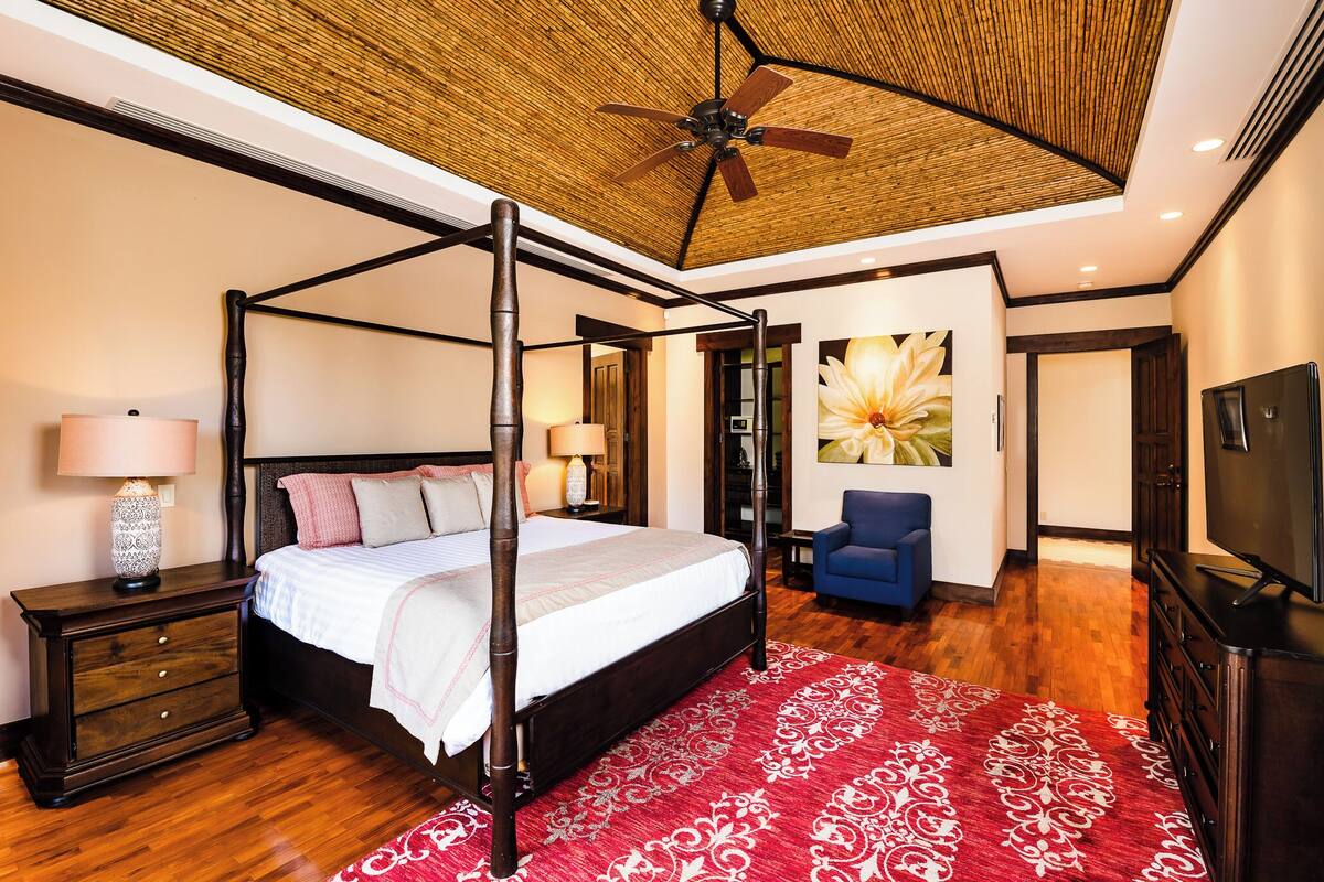 A four-poster bed with a red patterned rug, a ceiling fan, and a large floral painting on the wall.