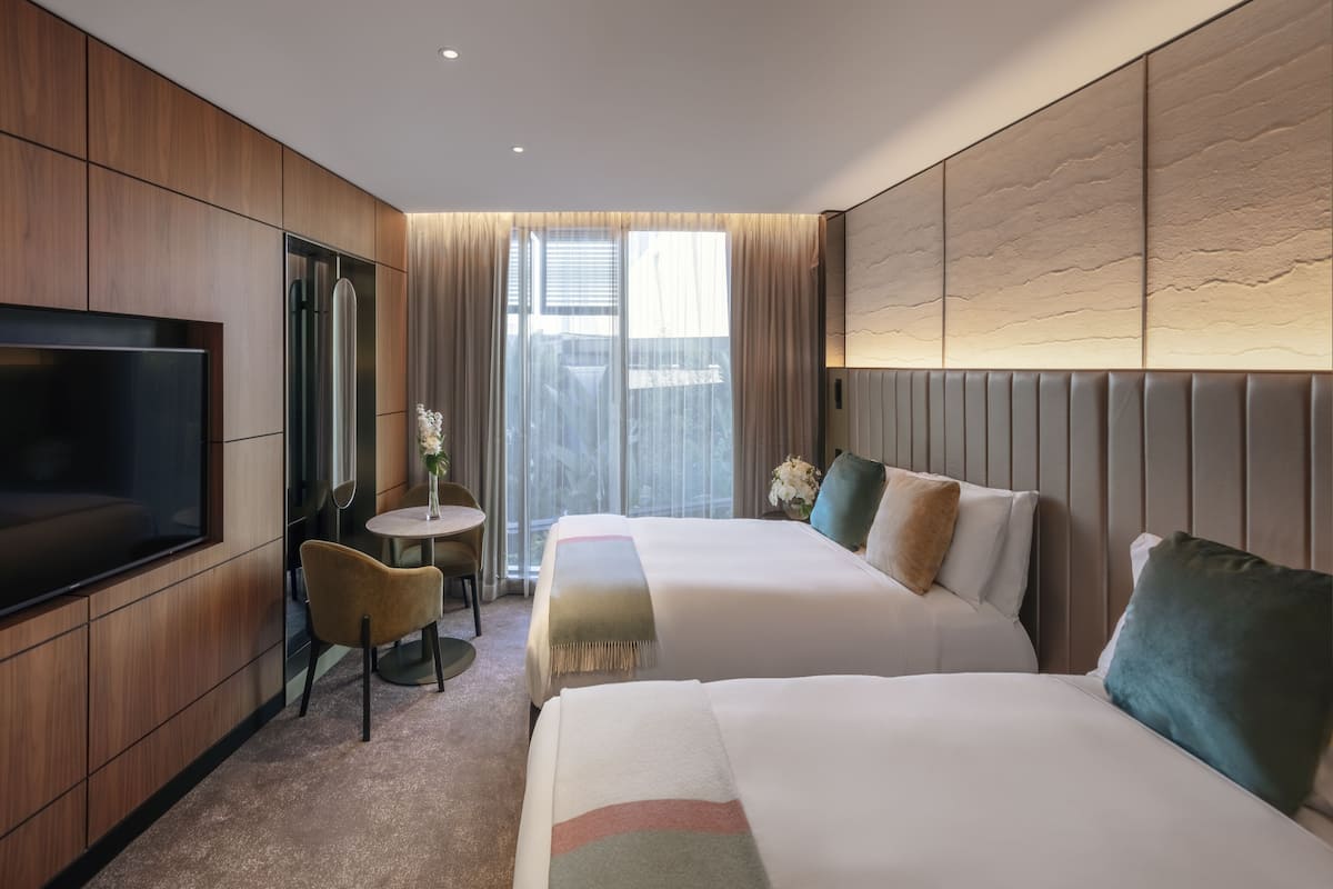 Una habitación de hotel con dos camas, un televisor, una mesita con una silla y una ventana con cortinas.
