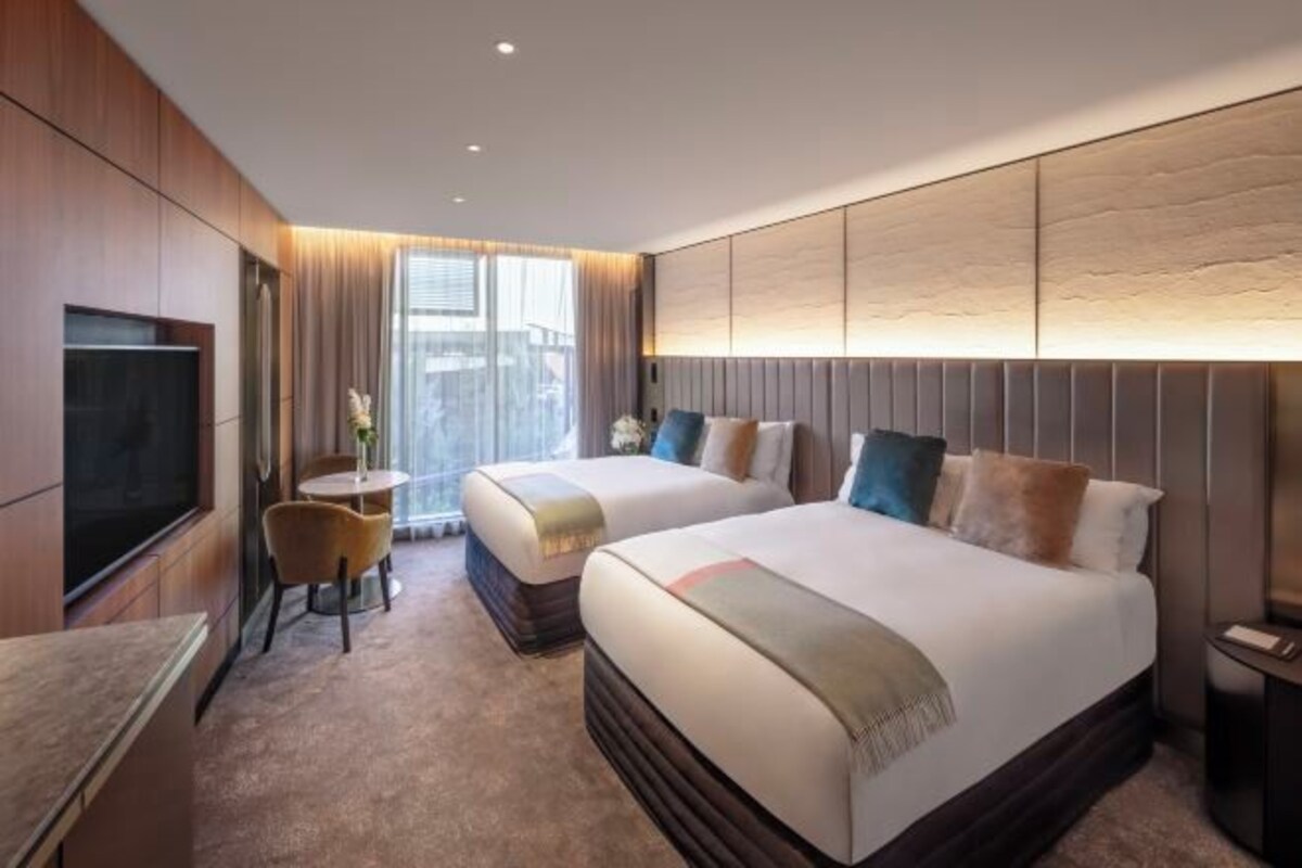 A hotel room with two beds, a TV, a small table with chairs, and a large window.
